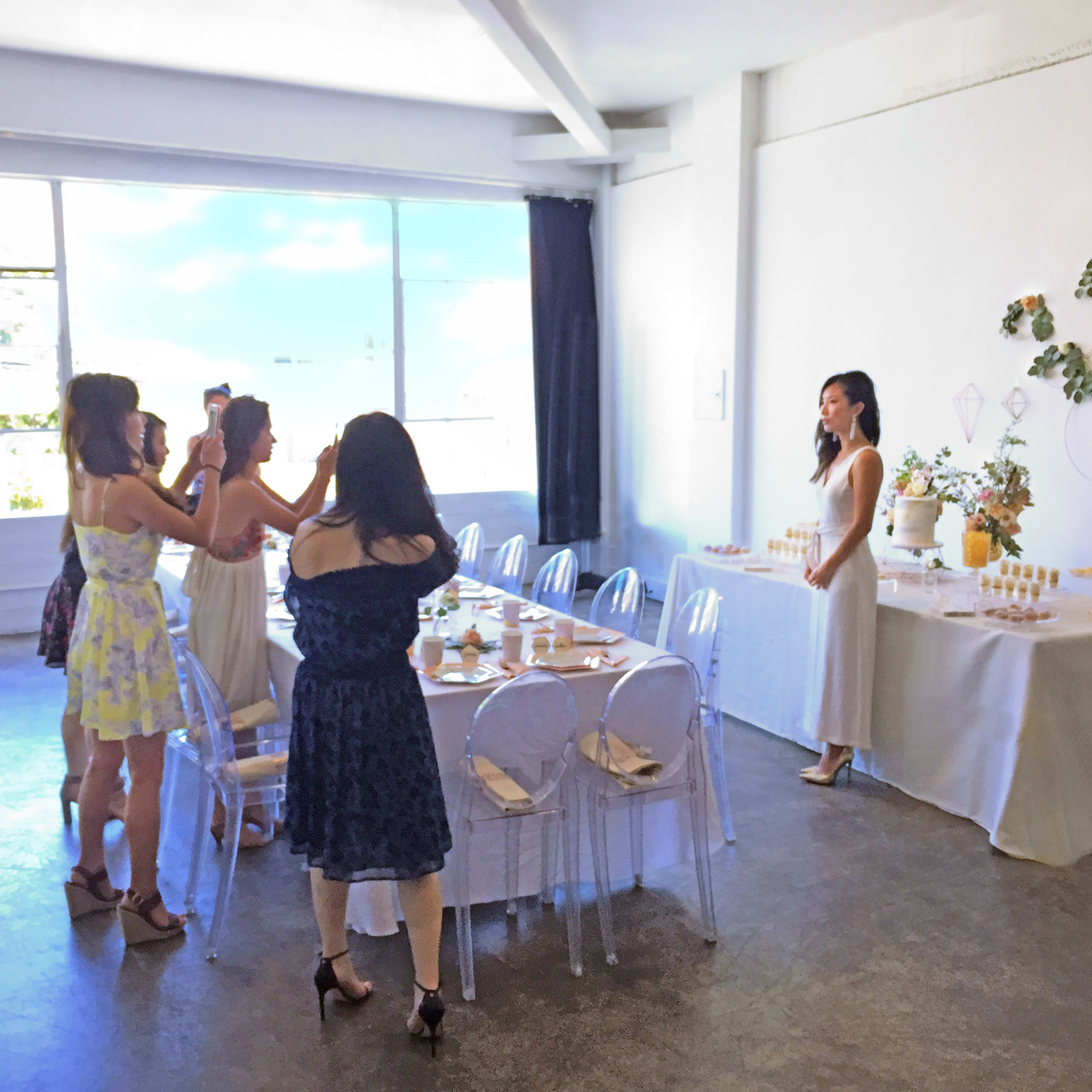 A group of women takes photos of a woman standing by a decorated table in a brightly lit room.