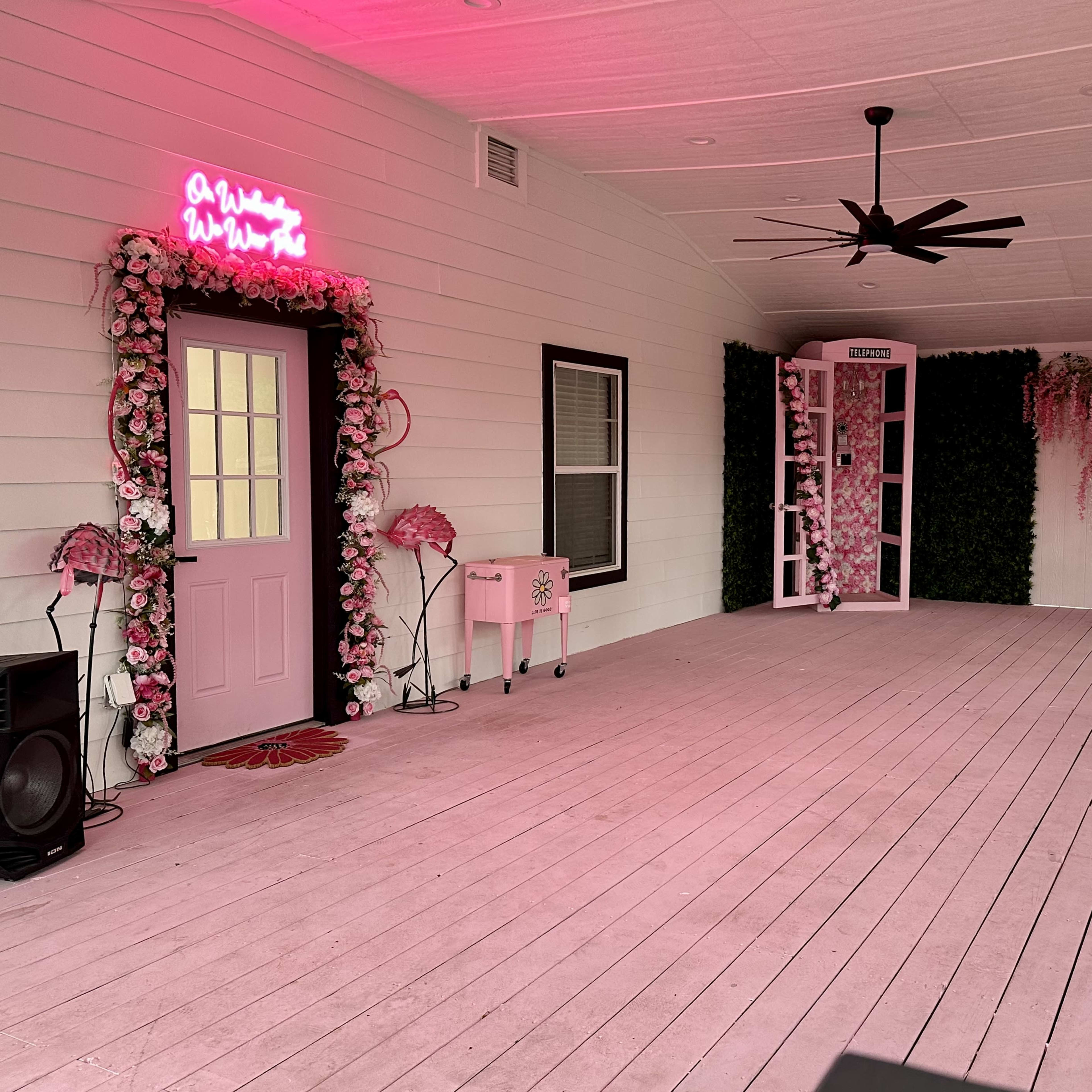 A decorated outdoor porch with pink walls, floral arrangements, seating, and a vintage phone booth.