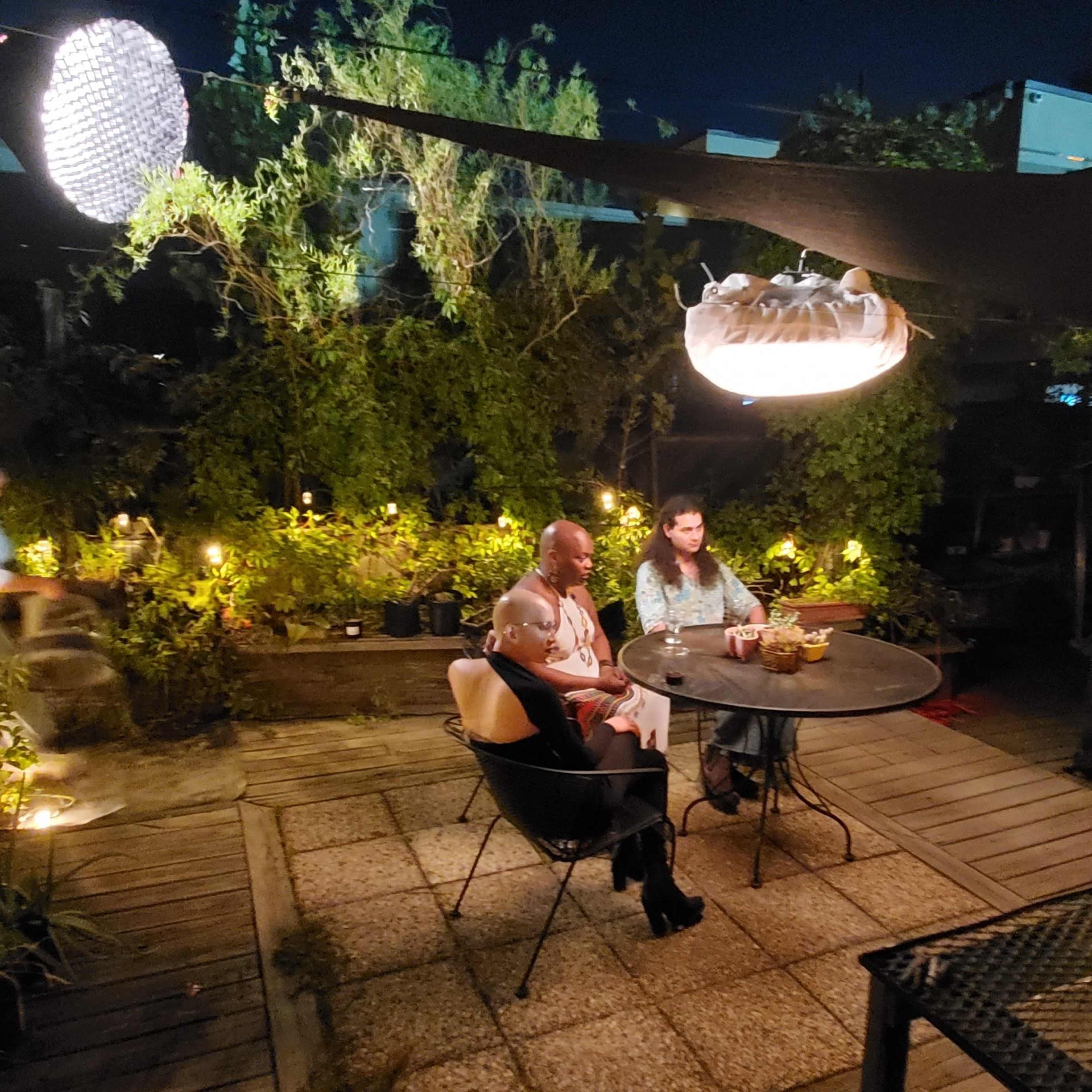 A night scene in a rooftop garden with several people sitting around a table surrounded by plants and soft lighting.