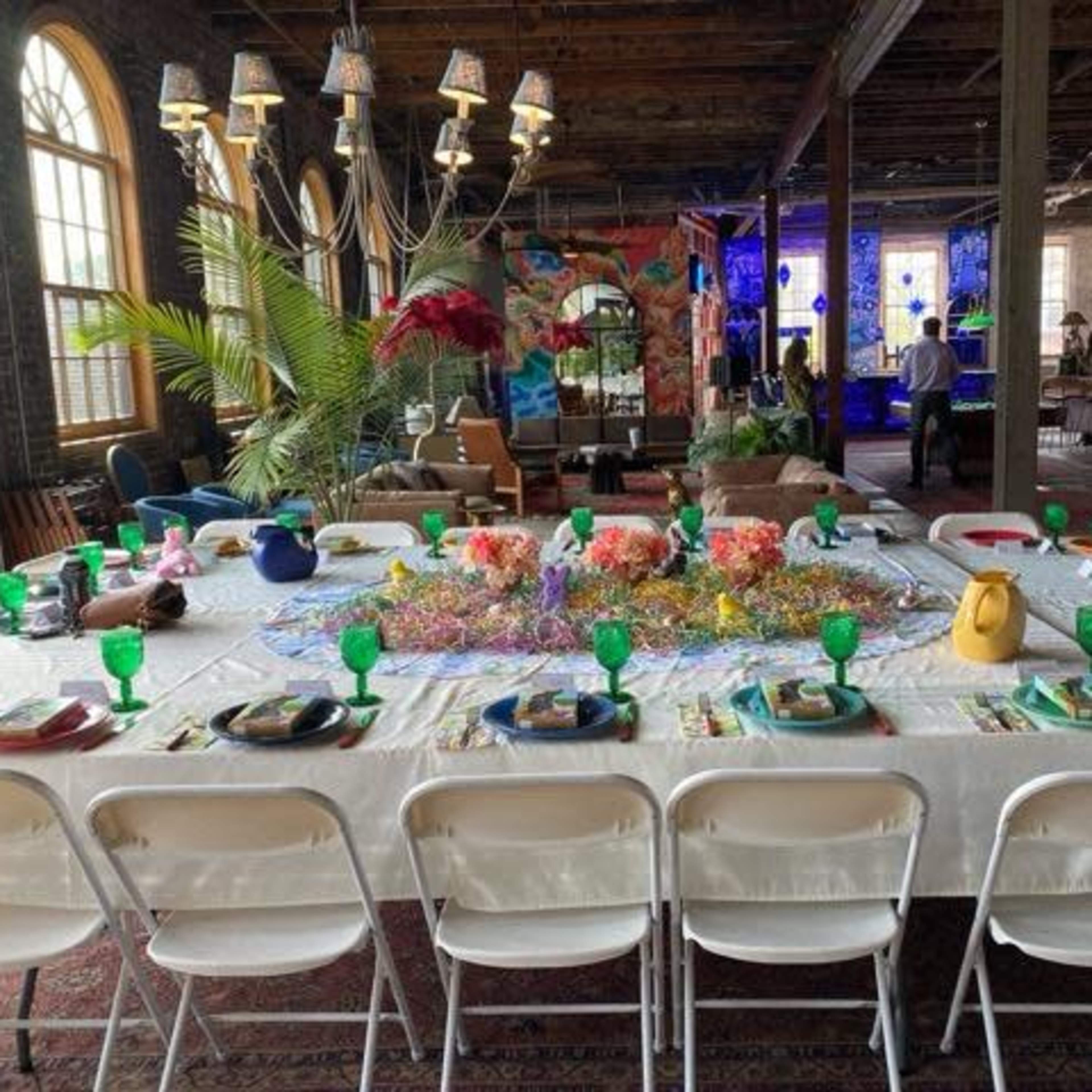 A long dining table is set with green glassware and colorful decorations in a spacious room with large windows and various lighting fixtures.