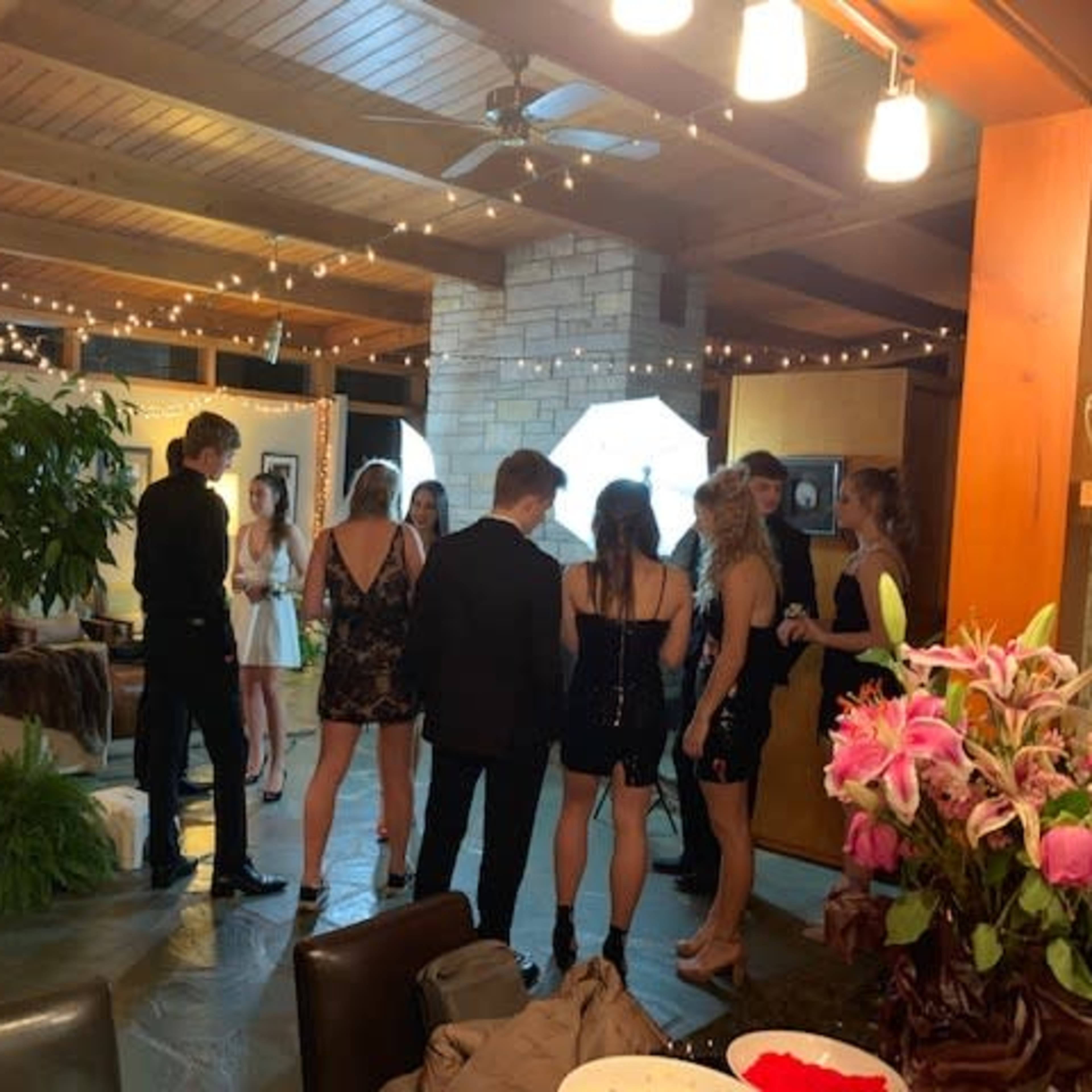 A group of people wearing formal attire gathers in a room decorated with string lights and indoor plants.