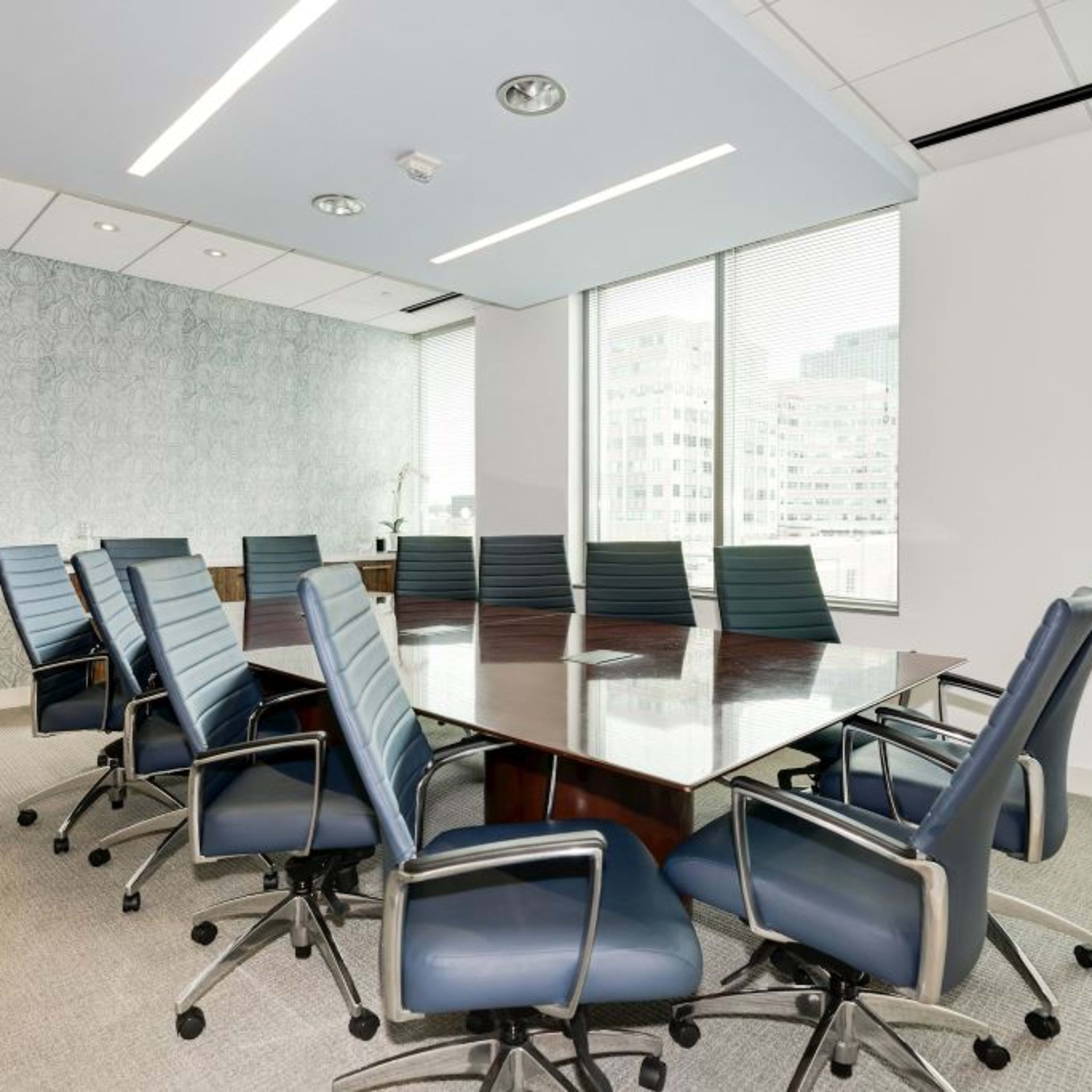 A conference room features a large wooden table surrounded by ergonomic office chairs and large windows allowing natural light.