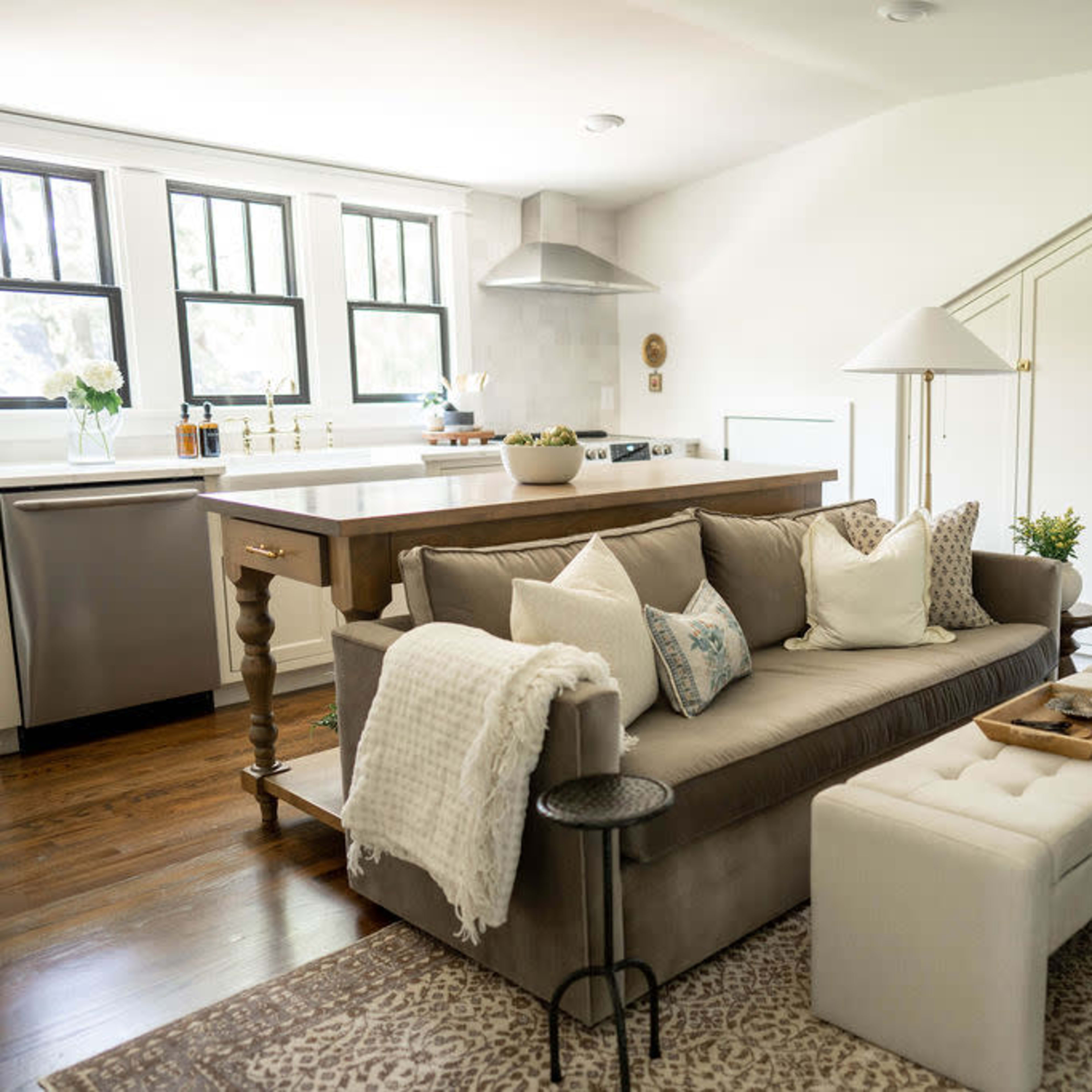 A light-filled living room featuring a beige sofa with decorative pillows, a white ottoman, and a kitchen area in the background with a wooden island.