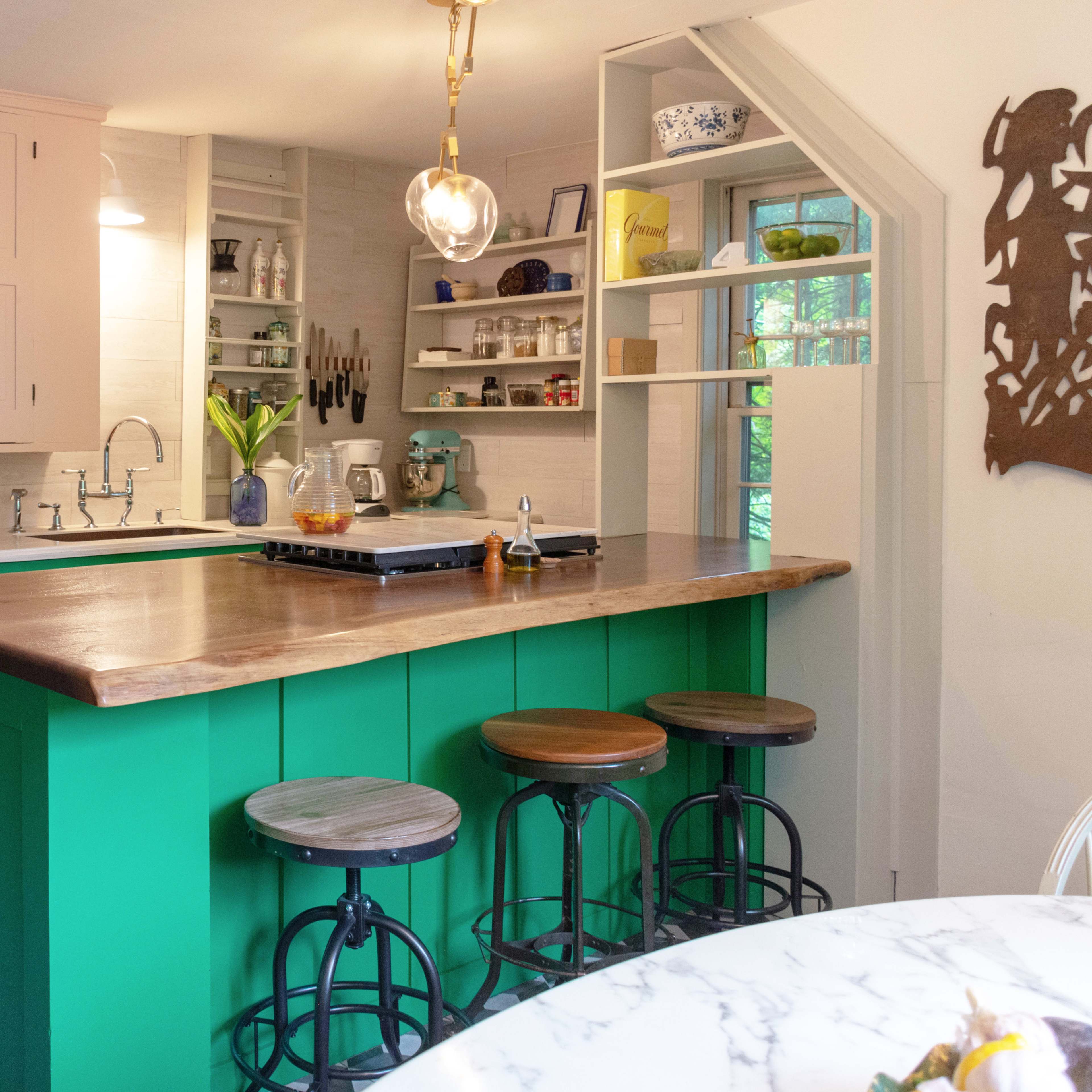 A kitchen with pink upper cabinets, a green island with a wooden countertop, and three bar stools, set against a tiled floor.