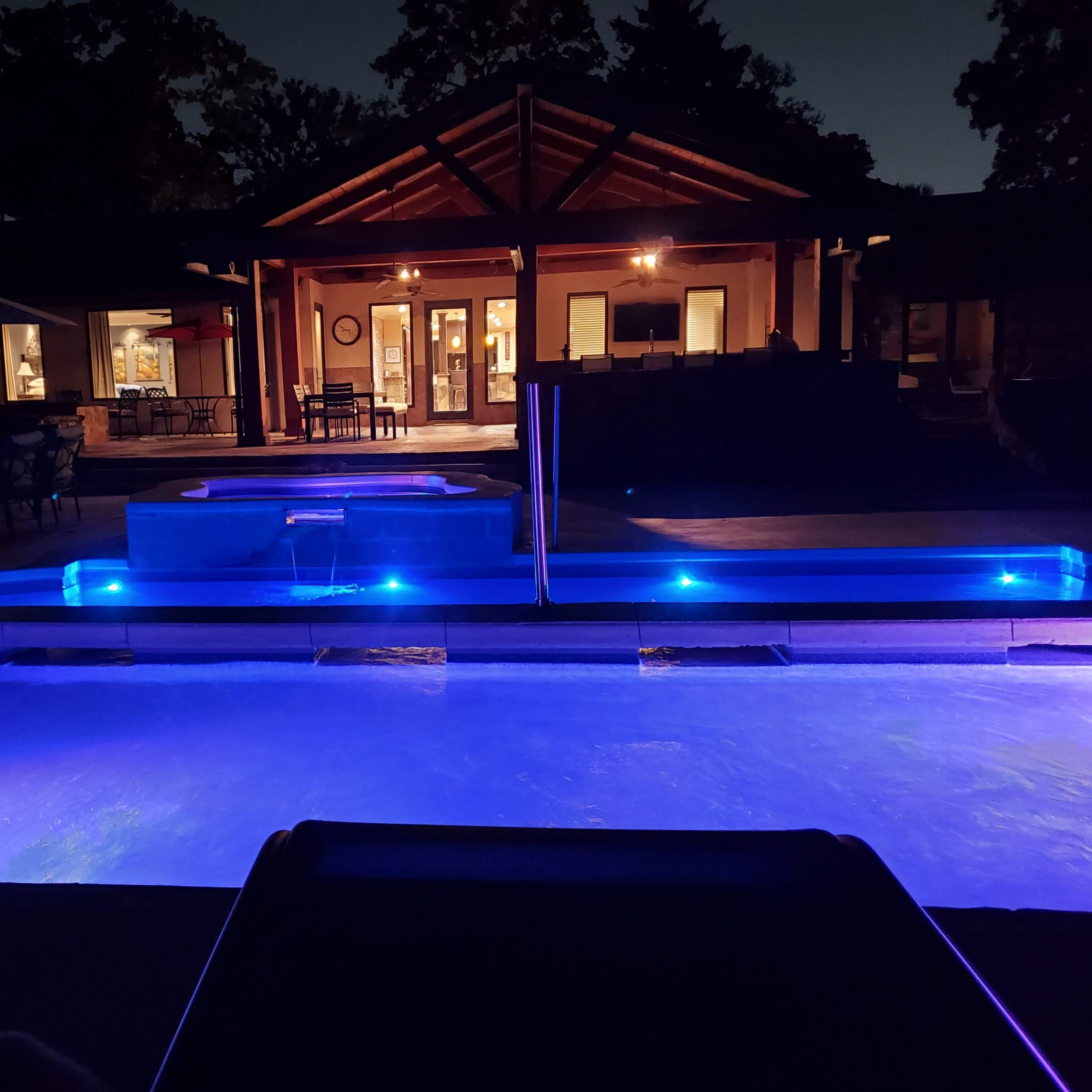 A lit pool area at night with a hot tub, a patio, and a house in the background.