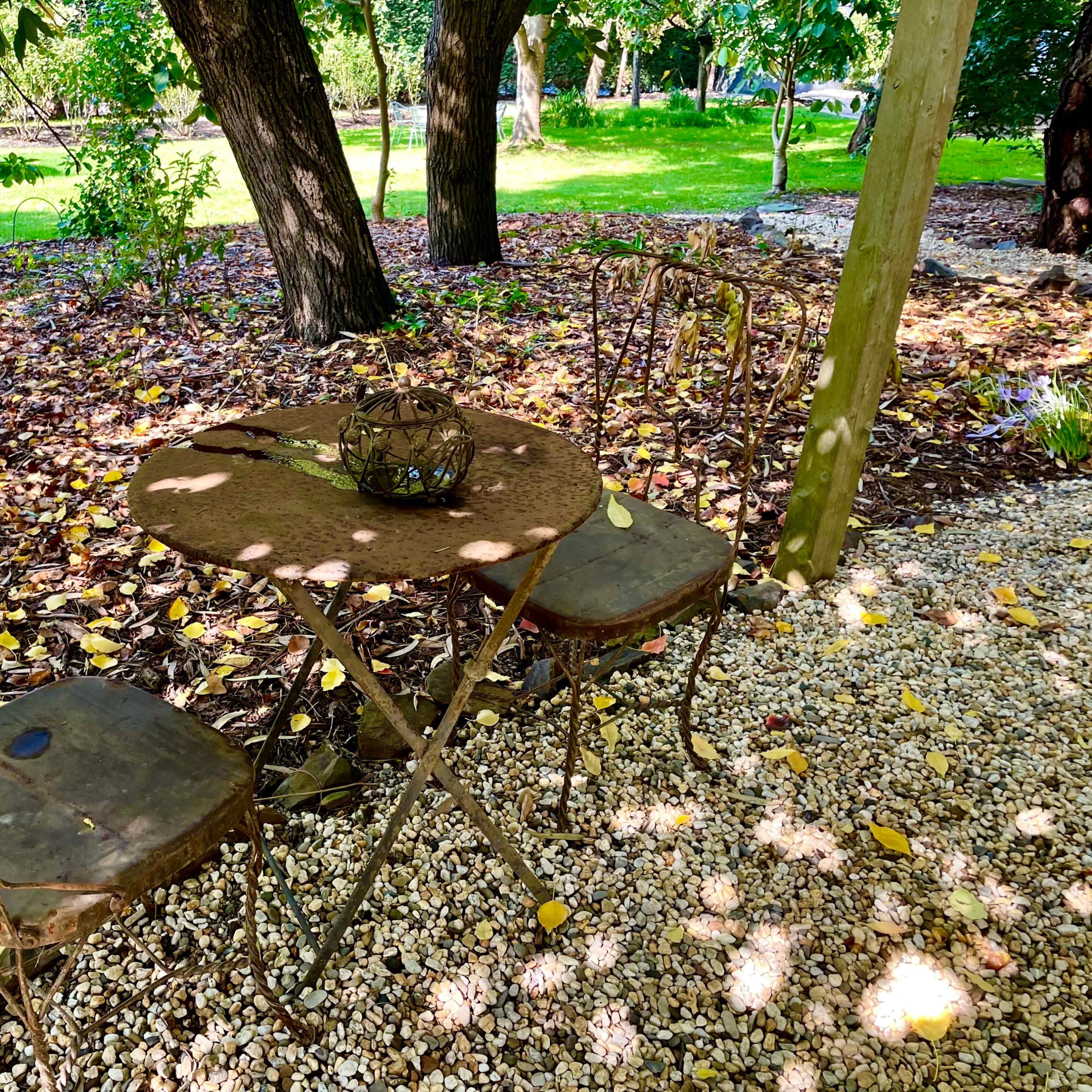 An outdoor table with three chairs sits on a gravel surface surrounded by trees and scattered leaves.
