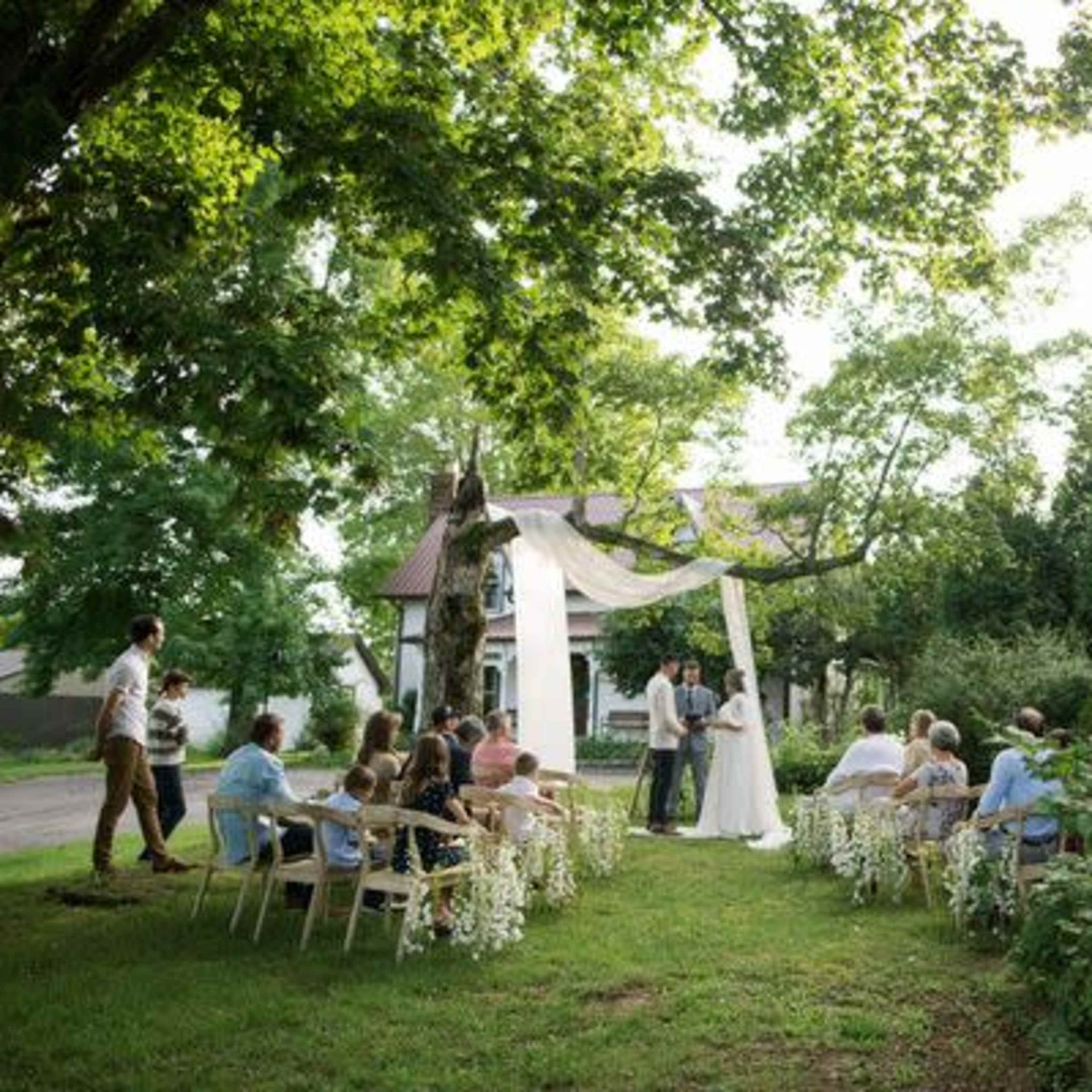 An outdoor wedding ceremony takes place under a draped canopy, surrounded by guests seated on chairs arranged in two rows.