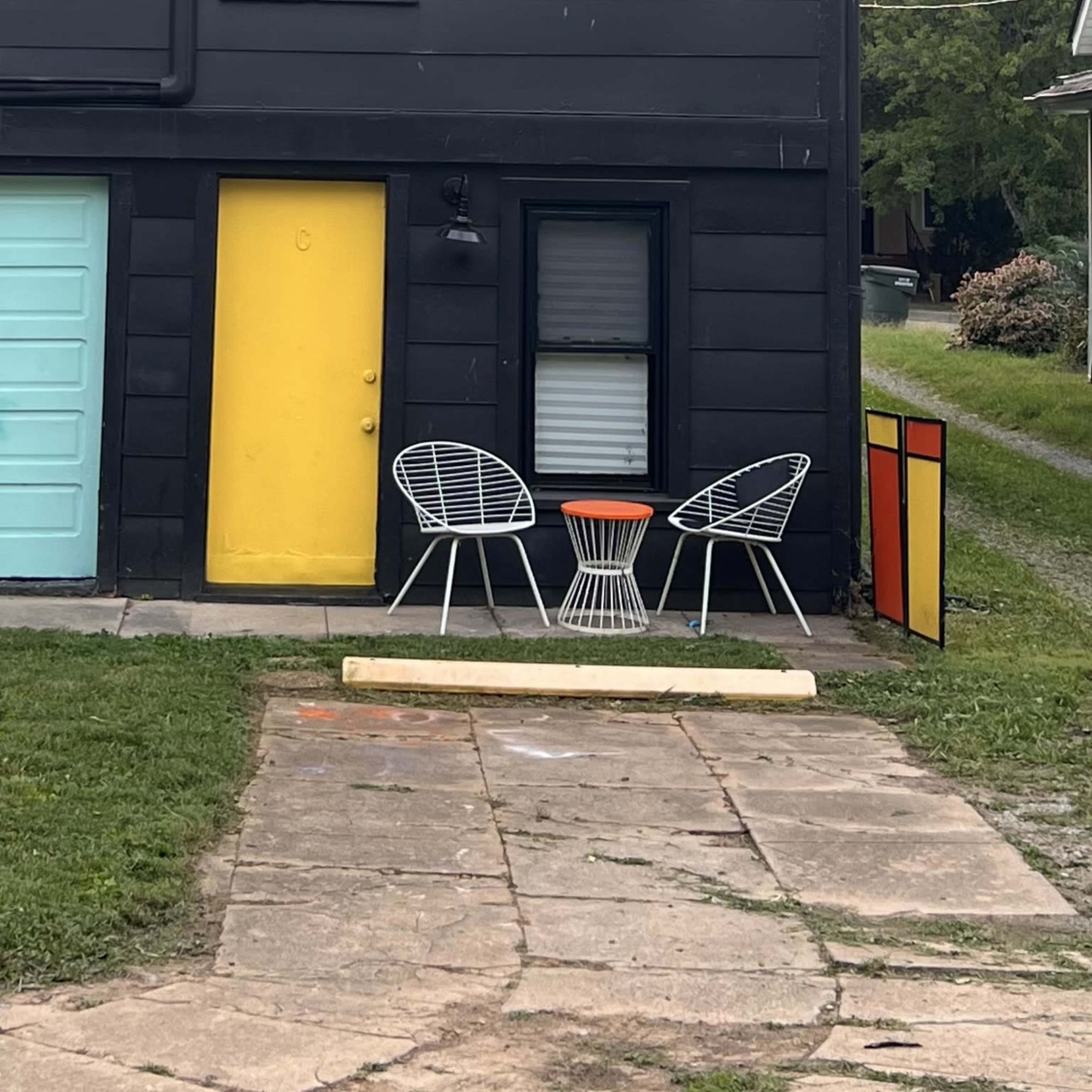 A small outdoor seating area with two white chairs and a round table, set against a black building featuring a yellow door and a colorful panel.