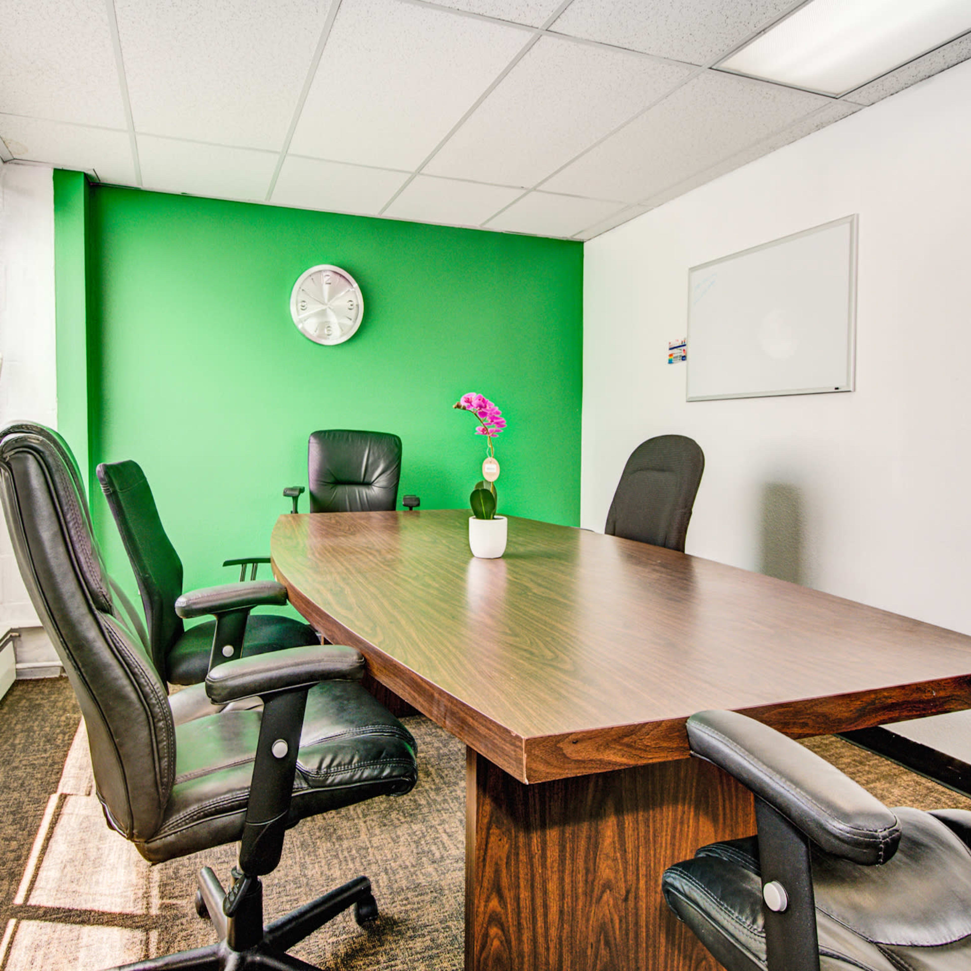 A conference room features a long wooden table surrounded by black office chairs, with a green accent wall and a clock mounted on the wall.