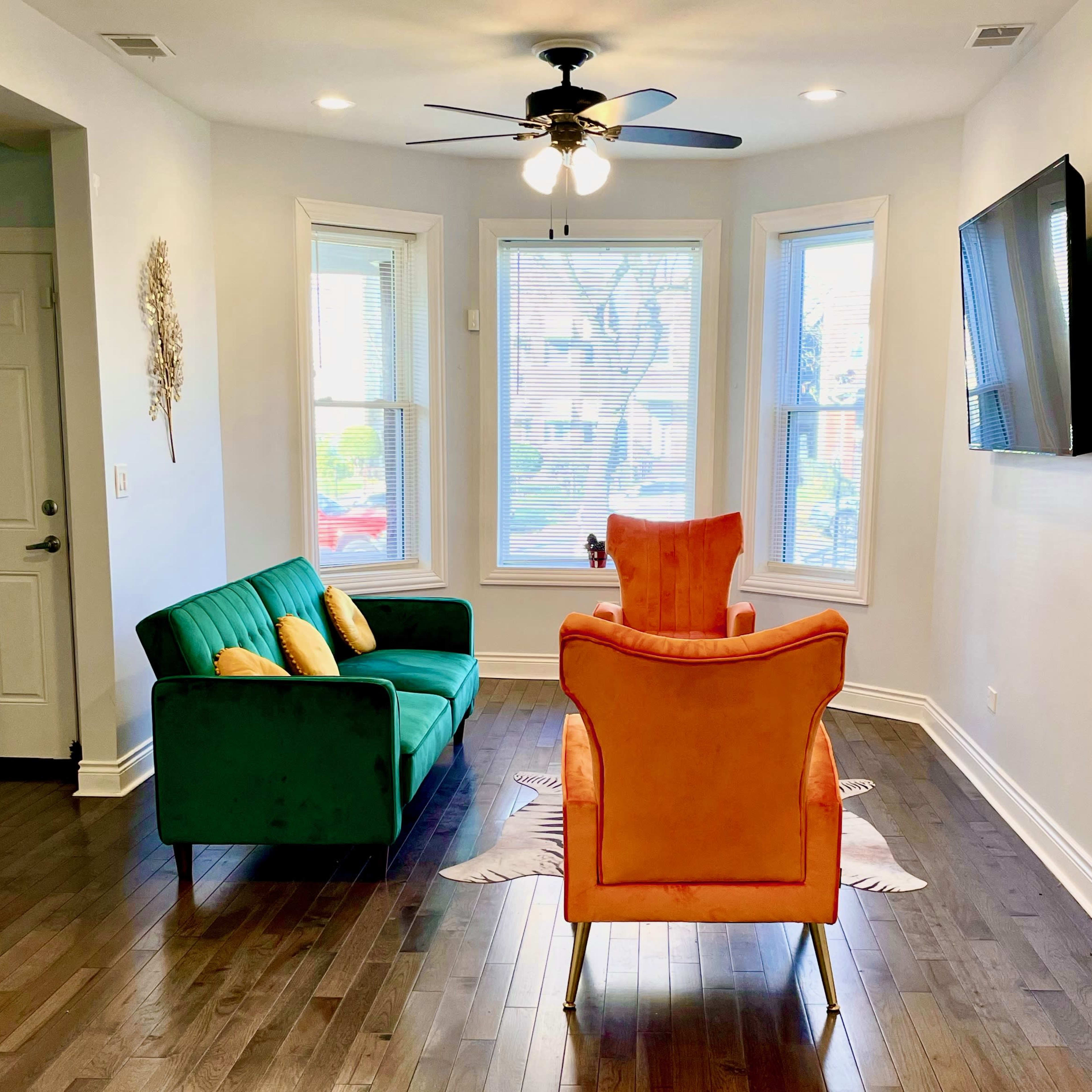 A living room with a green couch, an orange armchair, hardwood floors, large windows, and a mounted television on the wall.