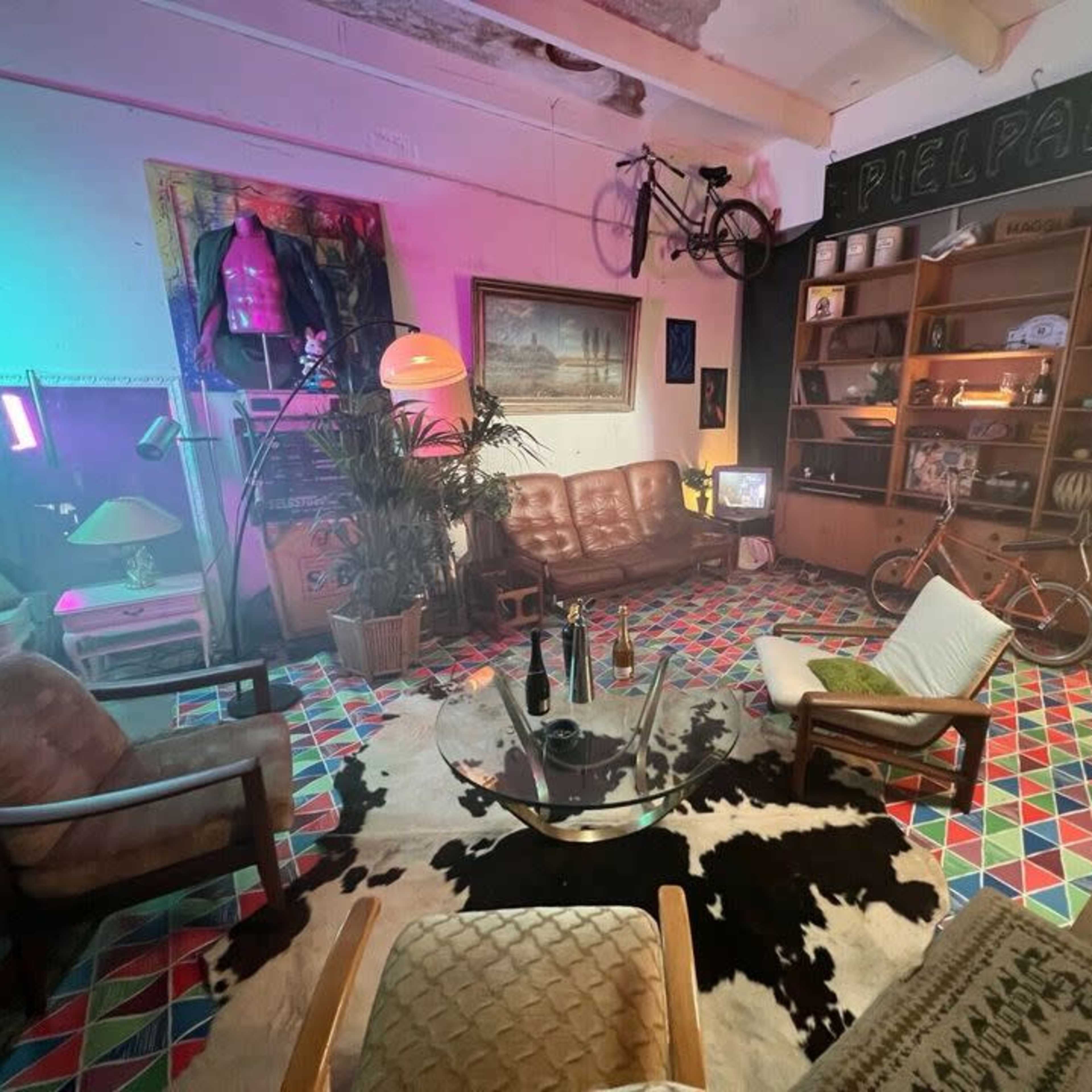 A retro-styled indoor living space with patterned flooring, vintage furniture, and a bicycle hanging from the ceiling, illuminated by colorful lights.