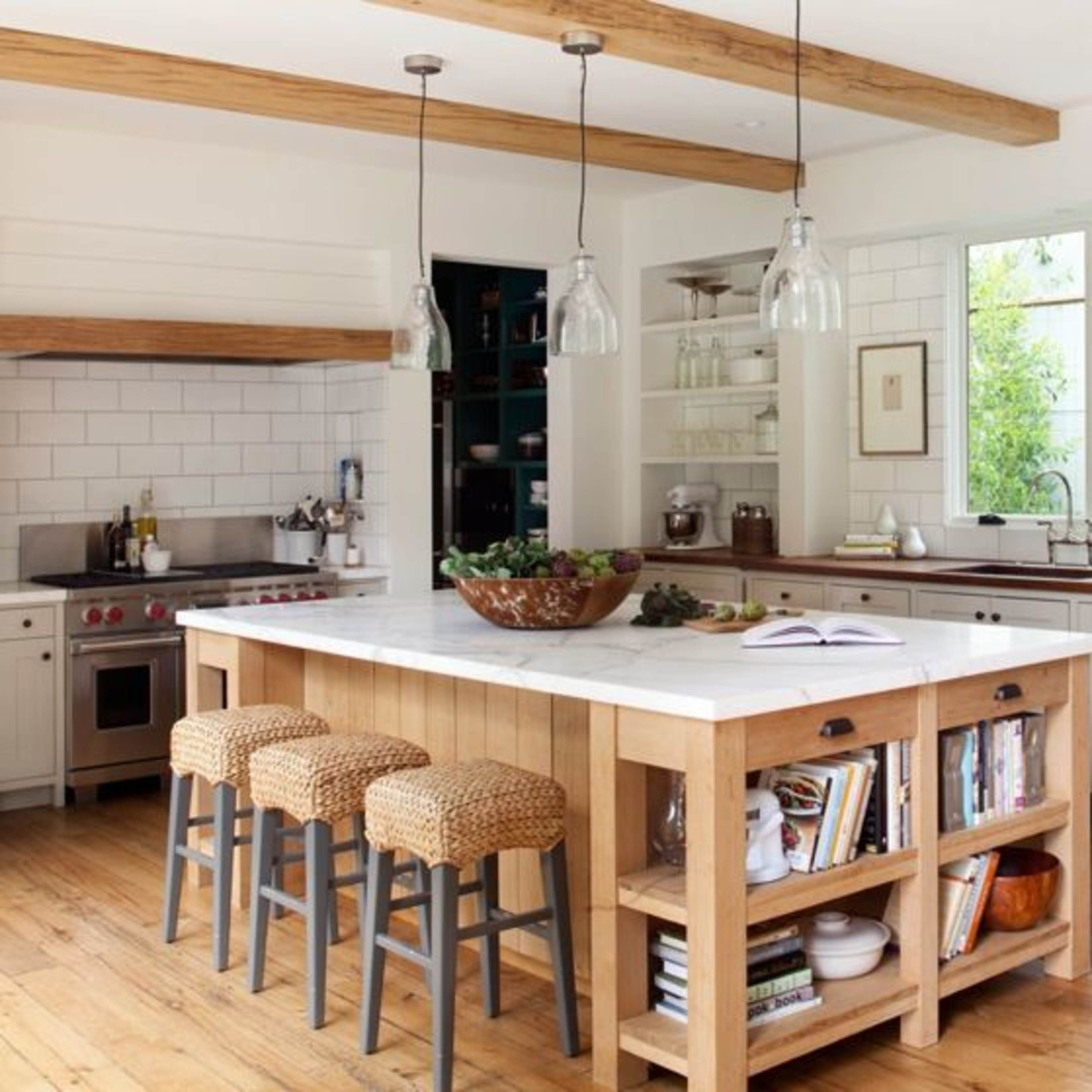 The kitchen features a large central island with a marble countertop, surrounded by woven bar stools, and is complemented by wooden beams and a window overlooking greenery.