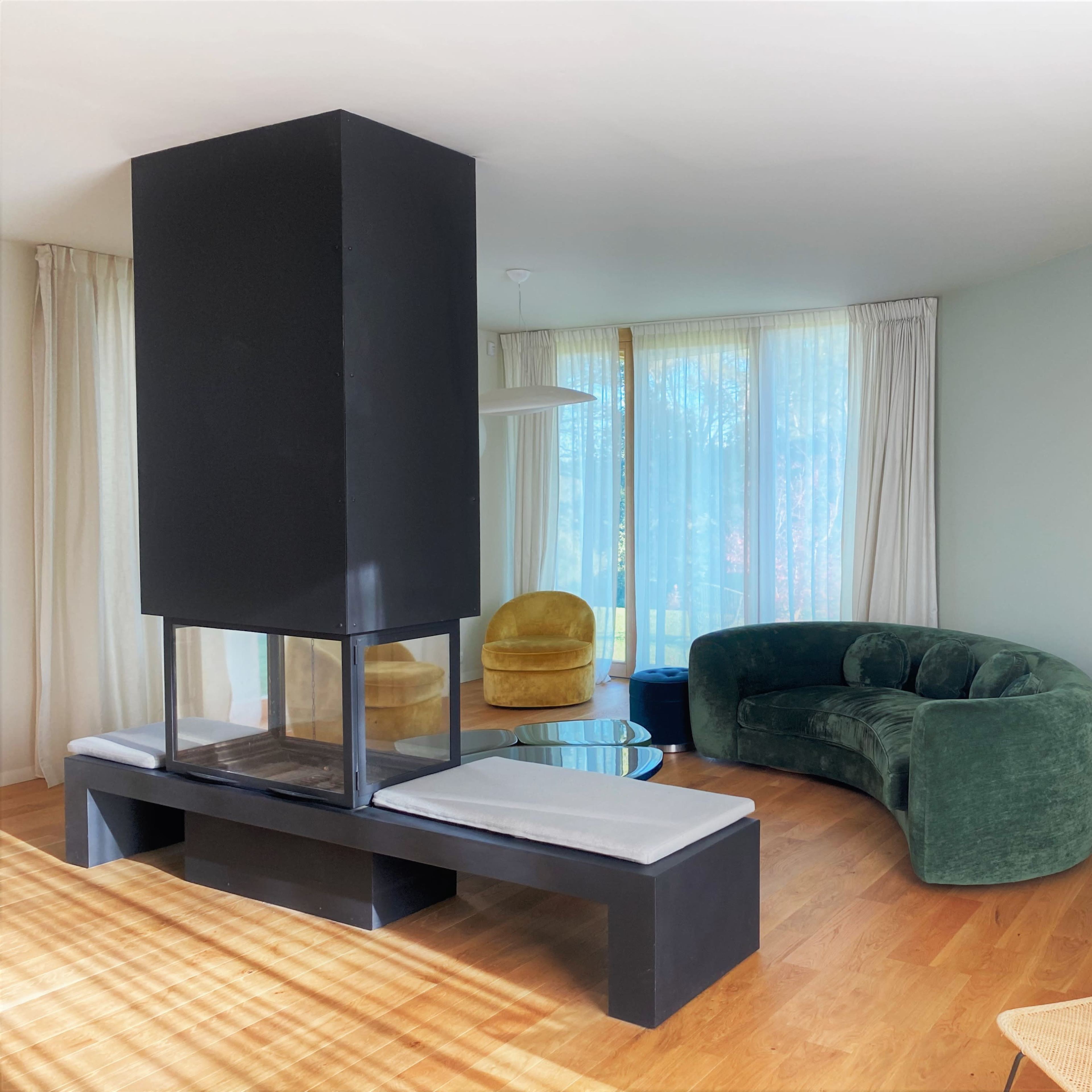A modern living room featuring a central black fireplace, a curved green sofa, a yellow armchair, and large windows letting in natural light.