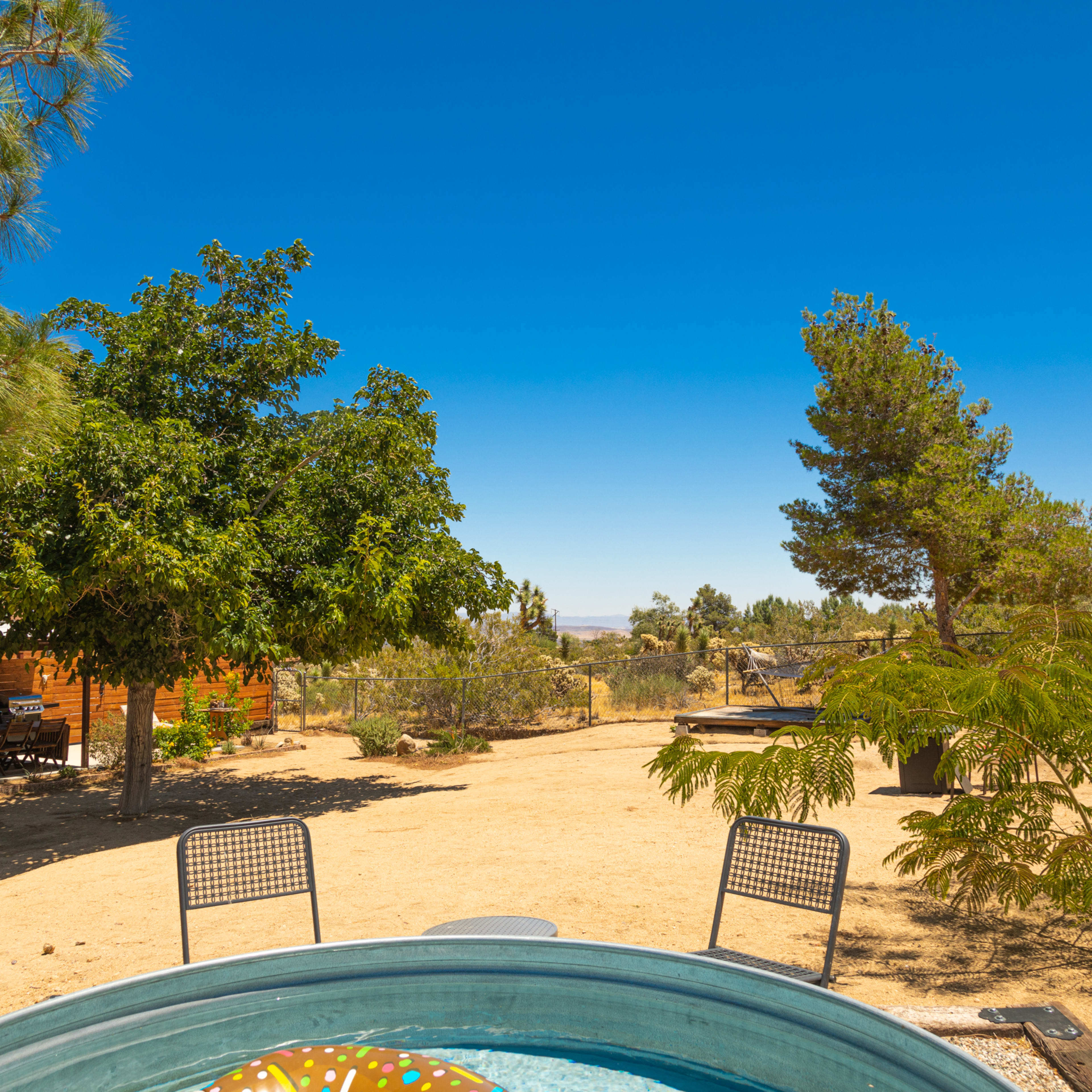 A hot tub sits surrounded by dry landscaping, with trees and a distant building visible under a clear blue sky.