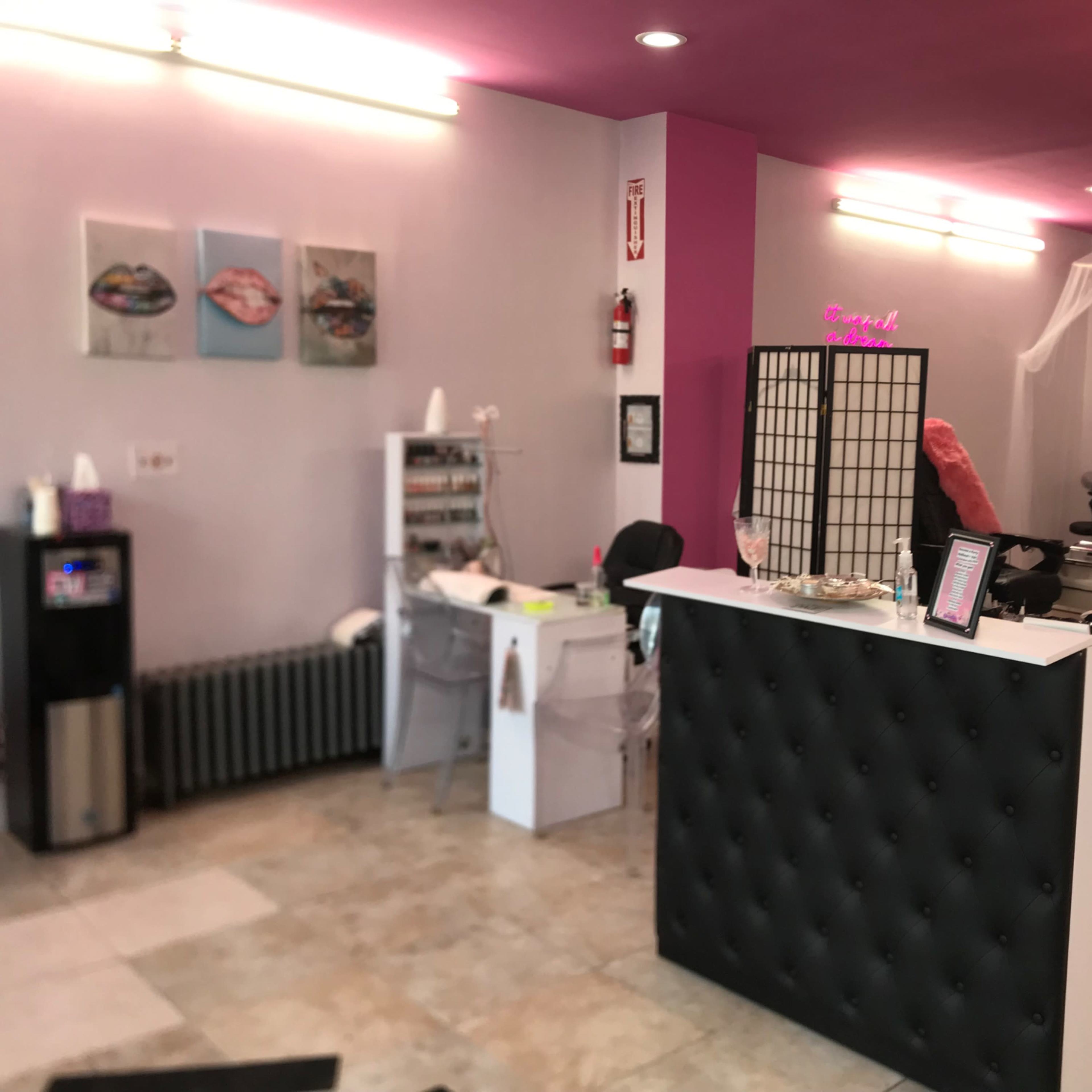 The interior of a beauty salon featuring a reception desk, nail service area, and a chic decor with pink and white accents.