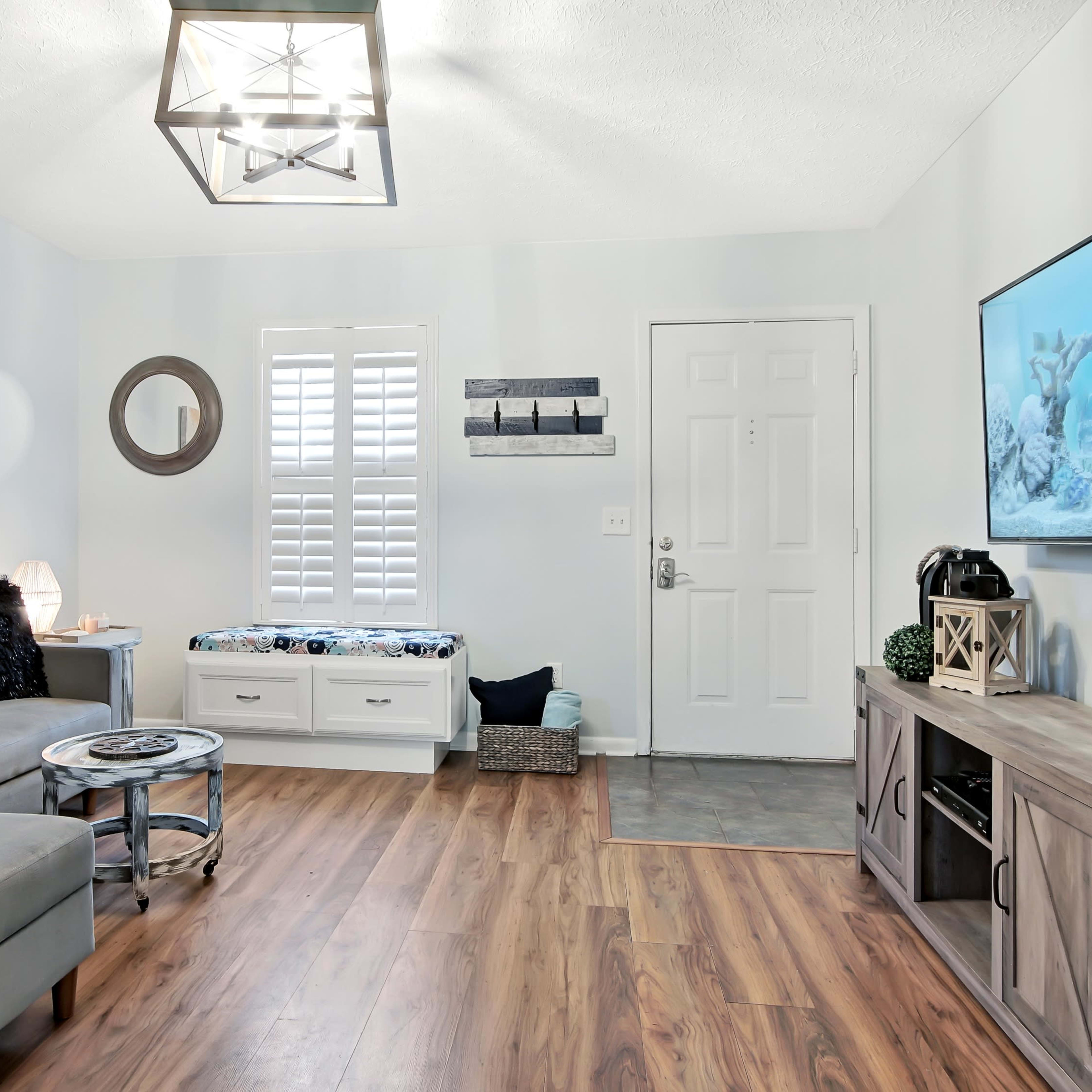 A living room with a gray sectional sofa, a wooden coffee table, a television mounted on the wall, and artwork hanging on the light-colored walls.