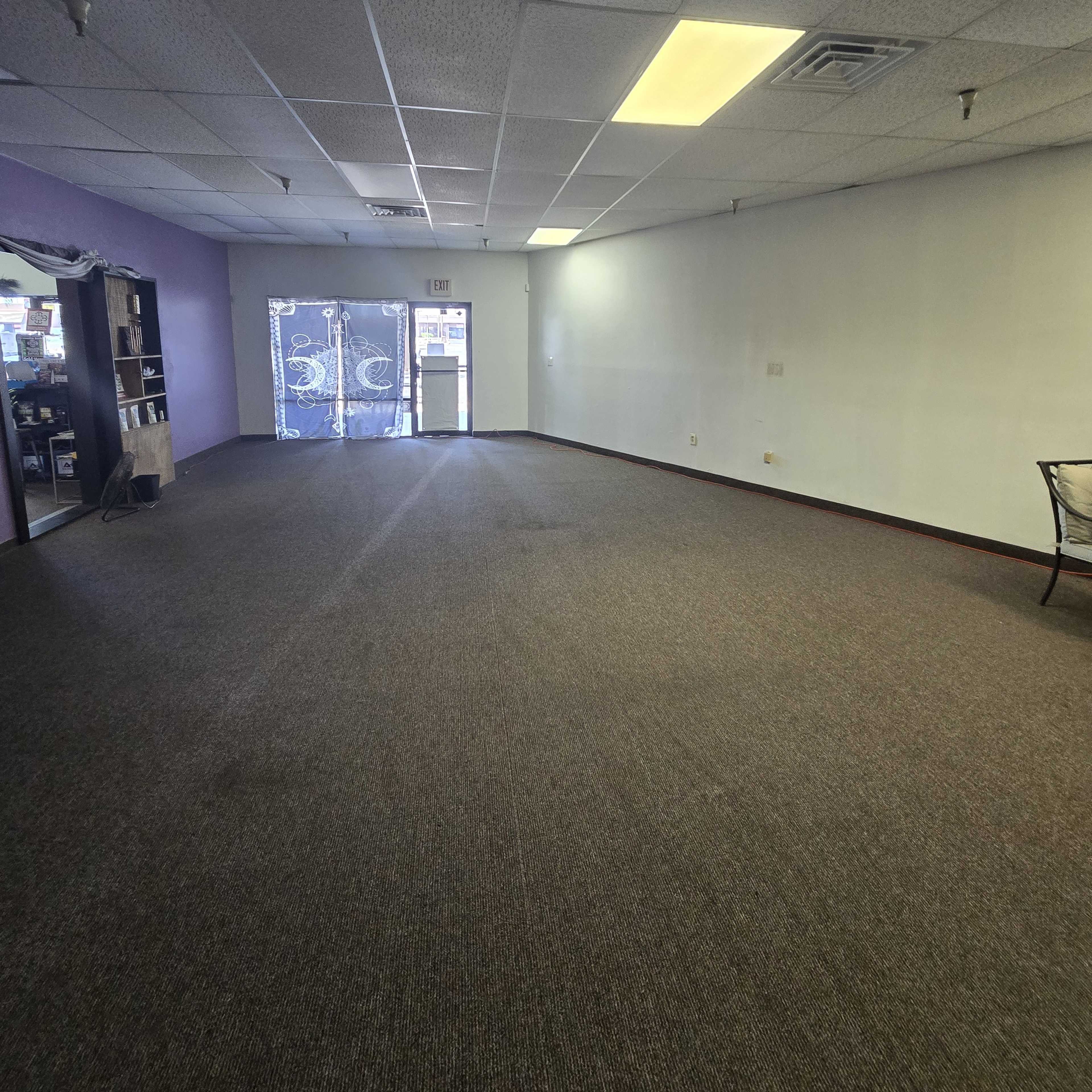 An empty commercial space with gray carpet and a purple accent wall, featuring a glass door leading outside.