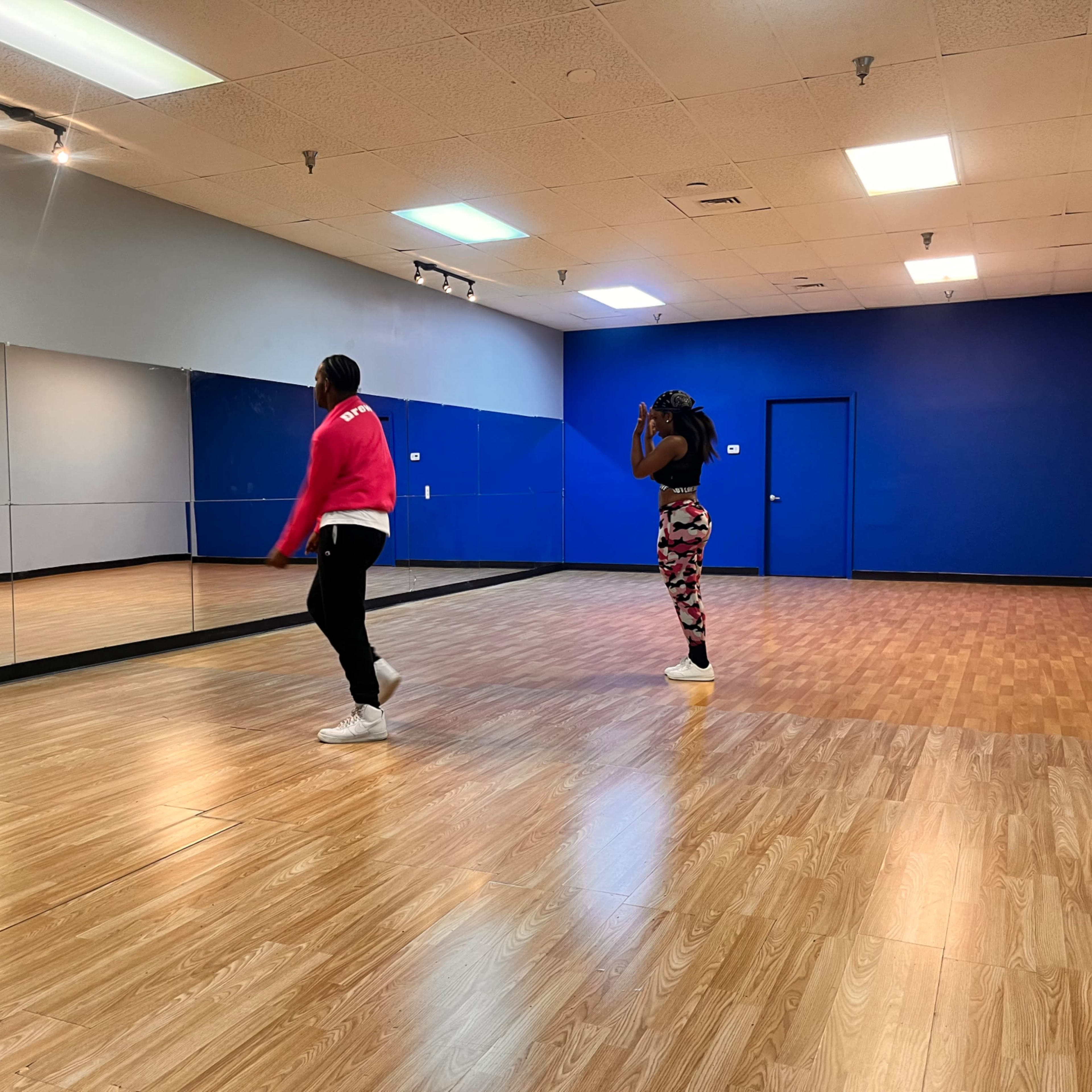 Two dancers practice in a mirrored dance studio with a blue accent wall and wooden flooring.