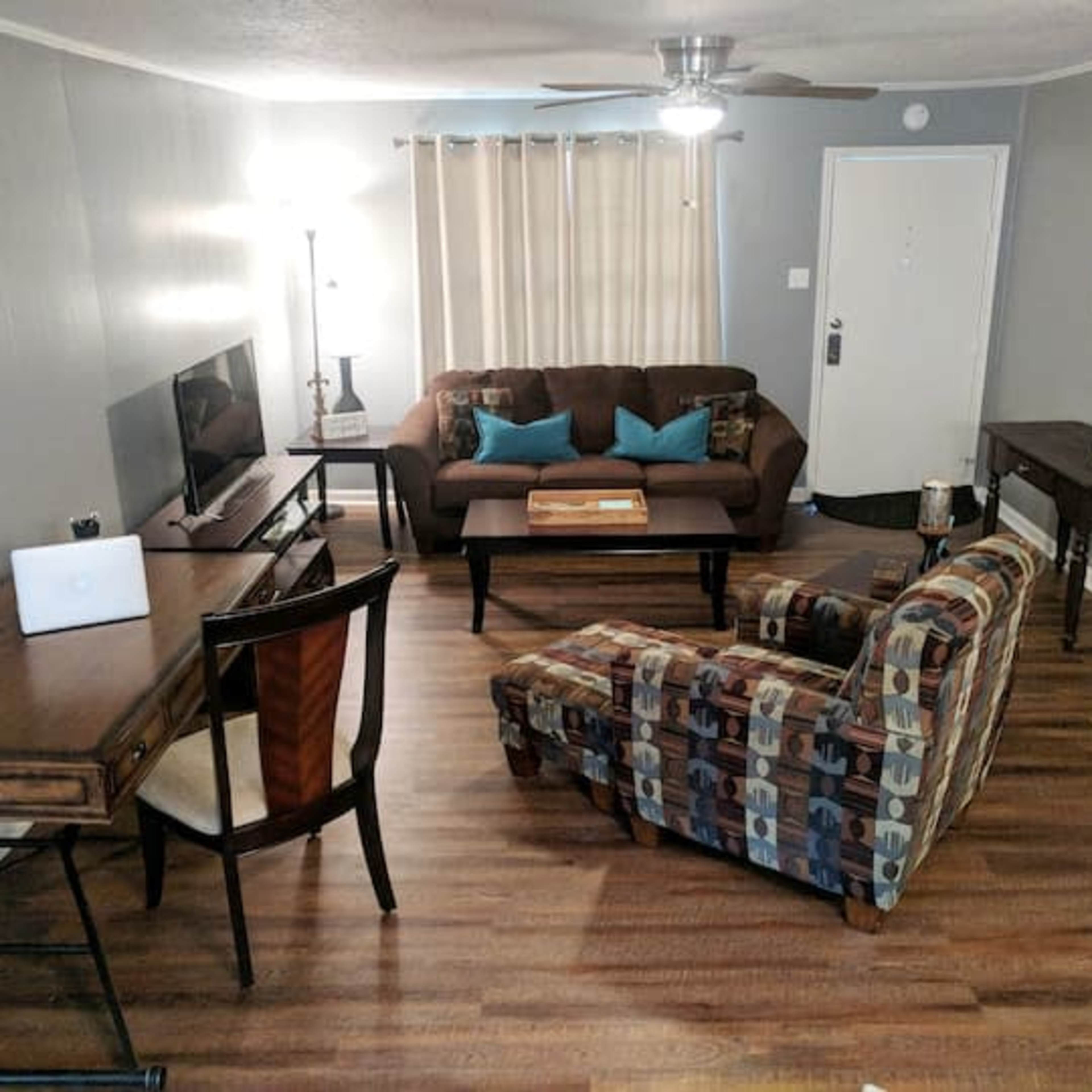 A living room featuring a brown couch, a chair with a patterned fabric, a wooden desk, and a television on a stand, all positioned around a central coffee table.