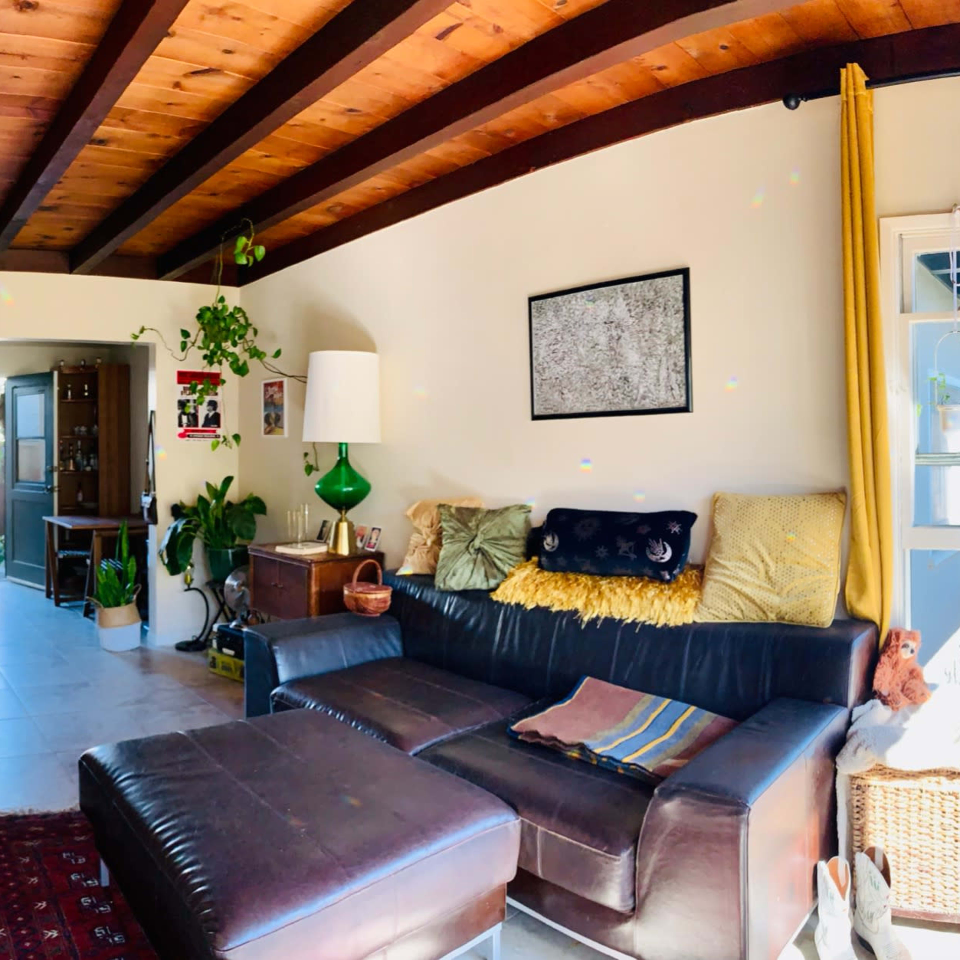A spacious living room with a sectional couch, plants, artwork on the walls, and large windows allowing natural light to enter.