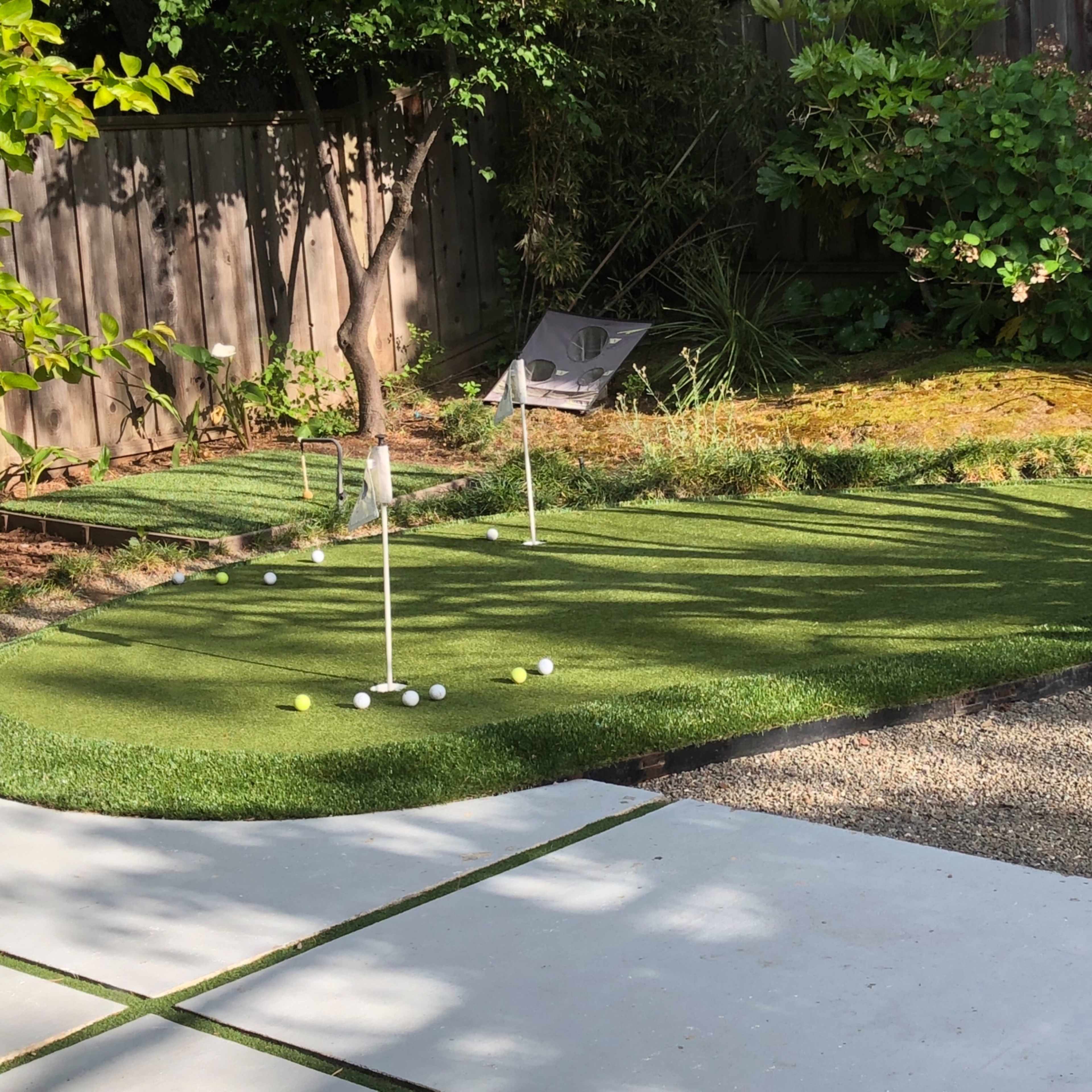 A backyard putting green with artificial turf, multiple holes, and a gravel pathway beside it.