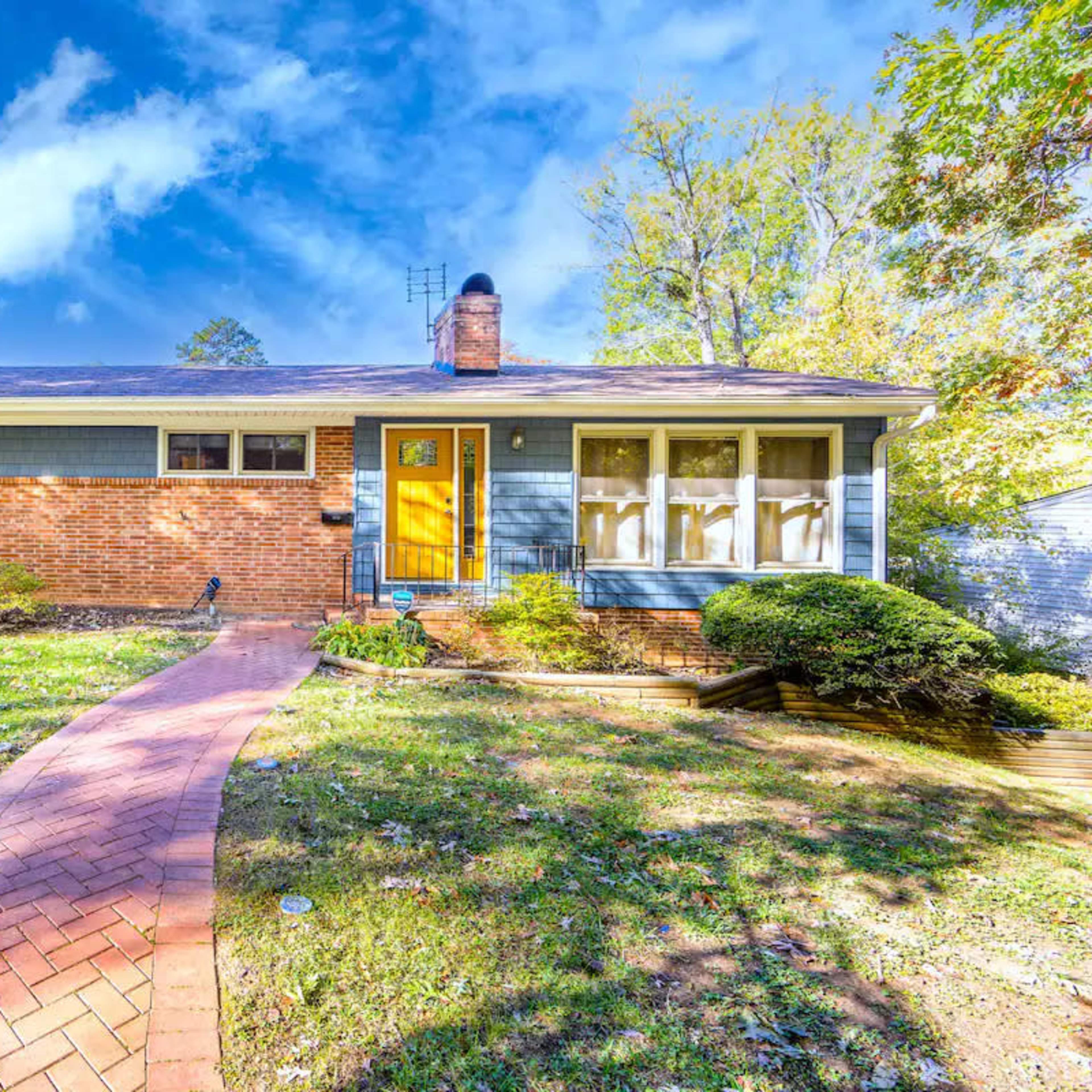 A brick ranch-style house with a blue exterior and a bright yellow door is surrounded by a landscaped yard and lined by a curved brick pathway.