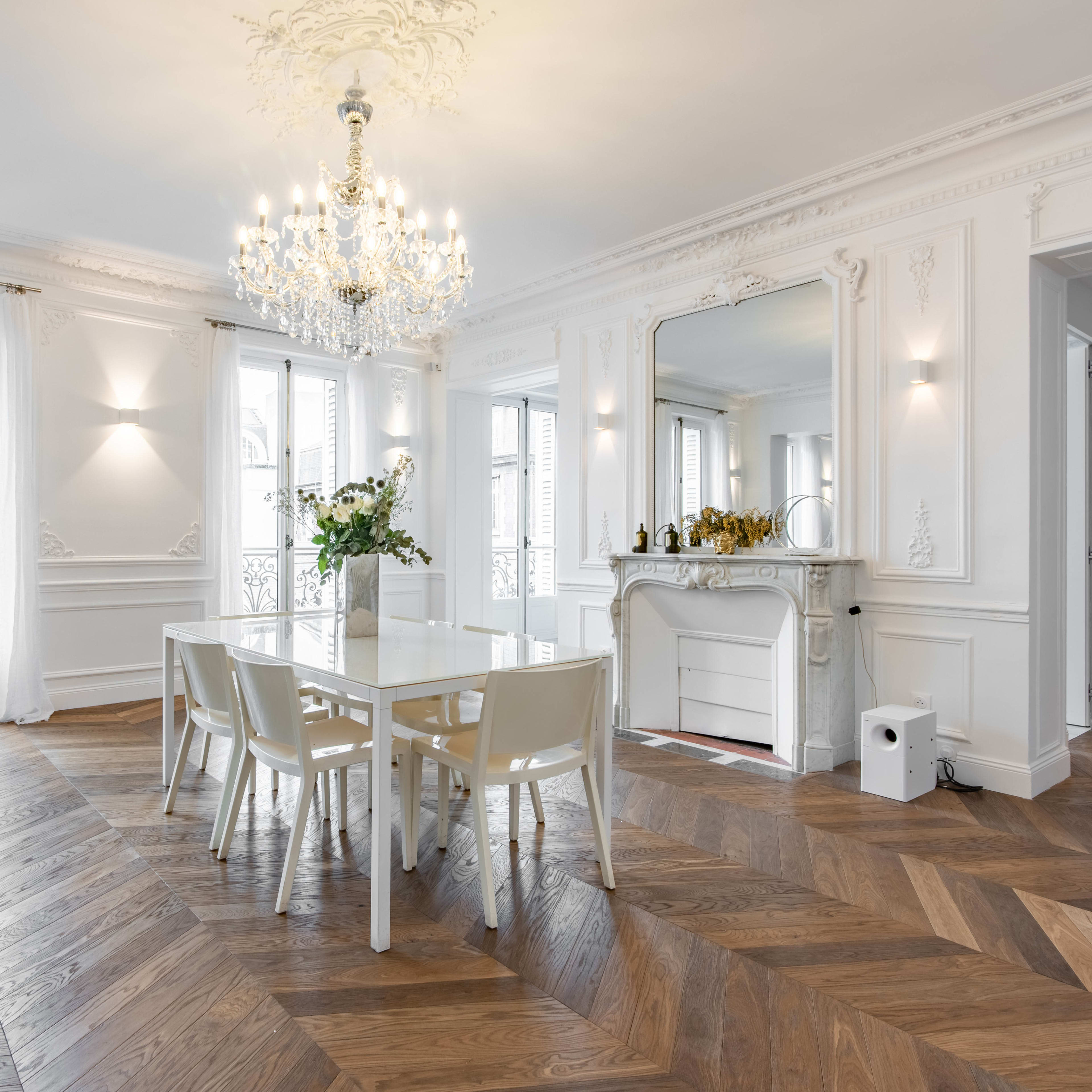 A bright dining area features a glass table surrounded by white chairs, with a chandelier overhead and ornate details on the walls.