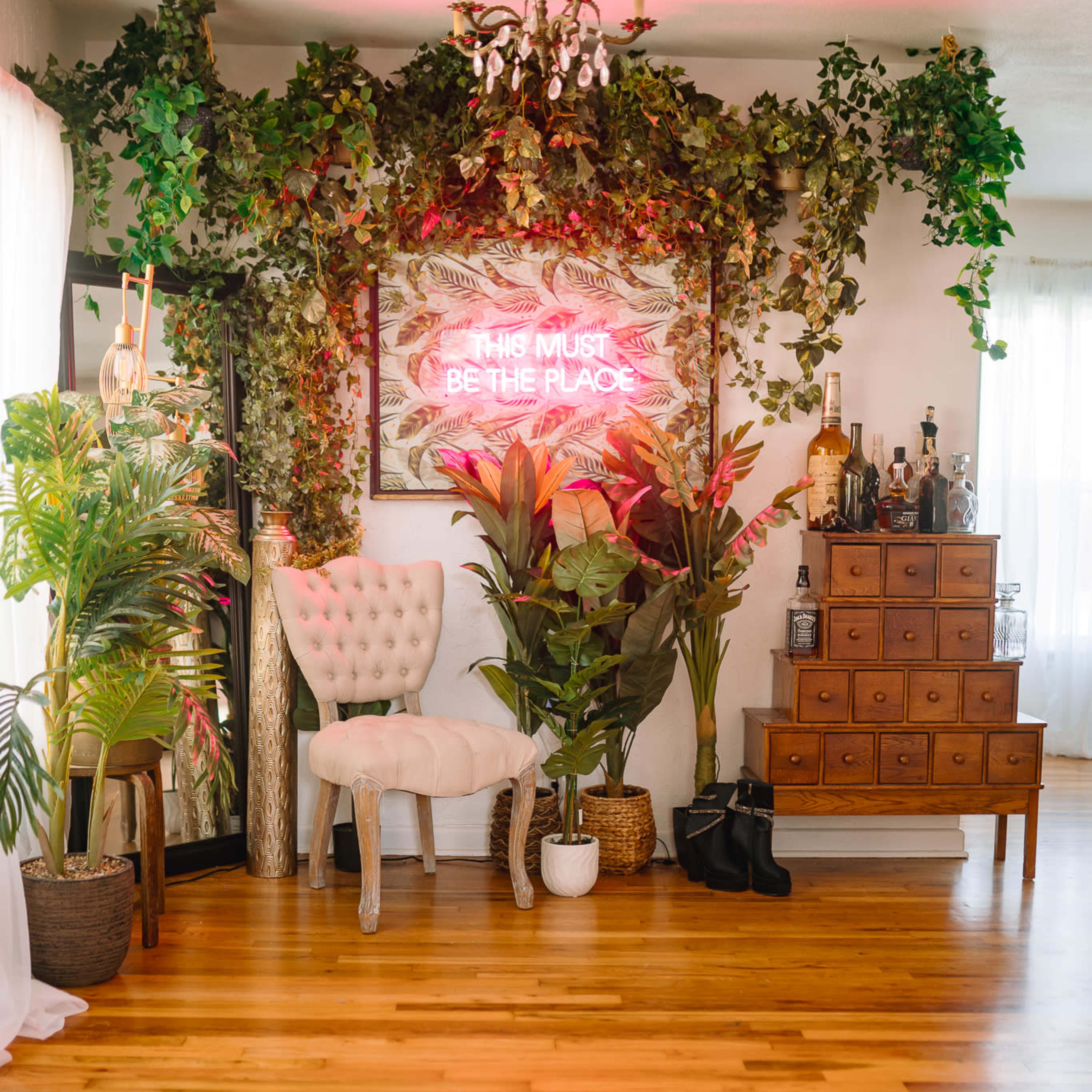A well-lit interior space features a decorative chair, lush greenery, a neon sign on the wall, and a wooden chest of drawers.