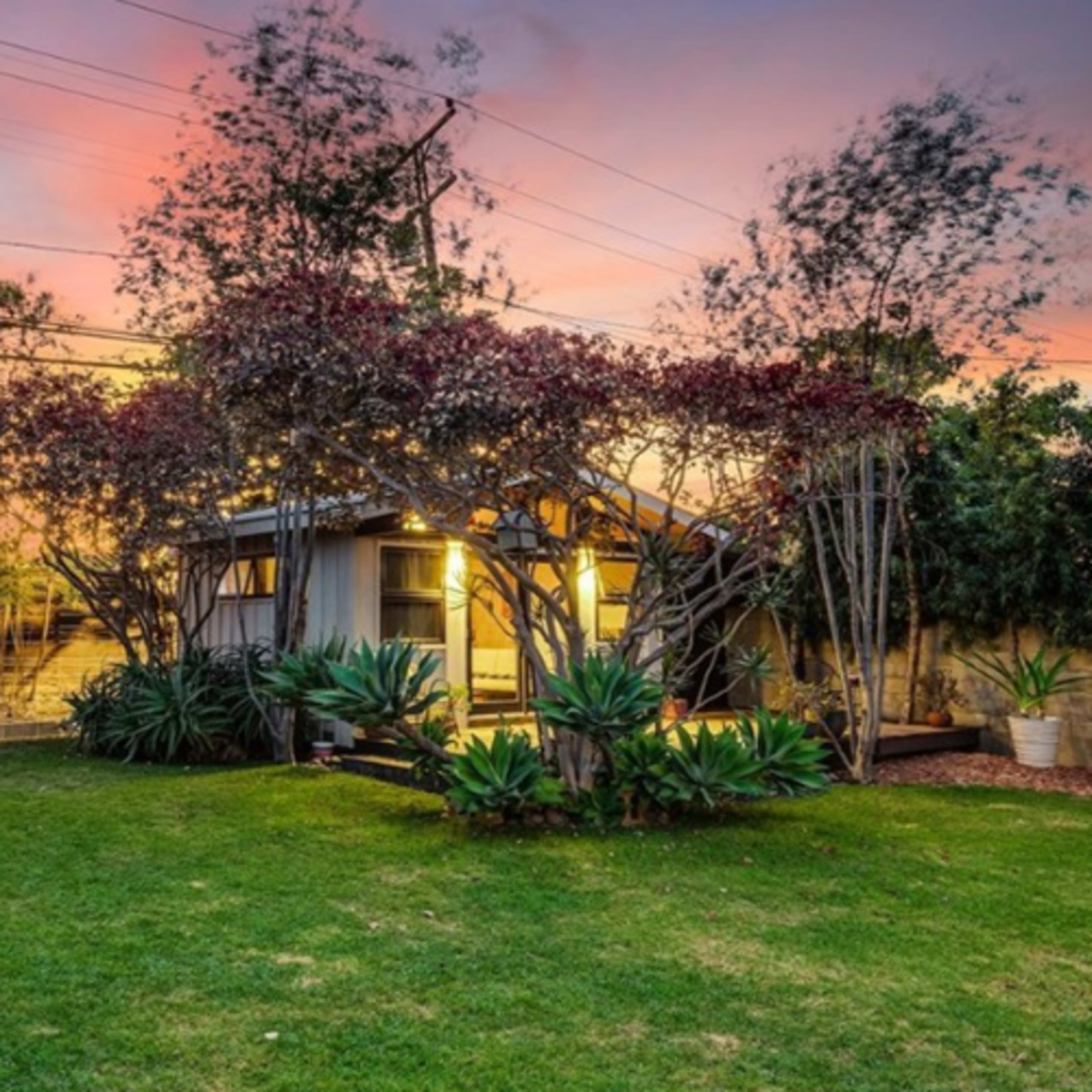 A house with a well-maintained lawn and surrounding trees is illuminated by evening light under a colorful sunset sky.