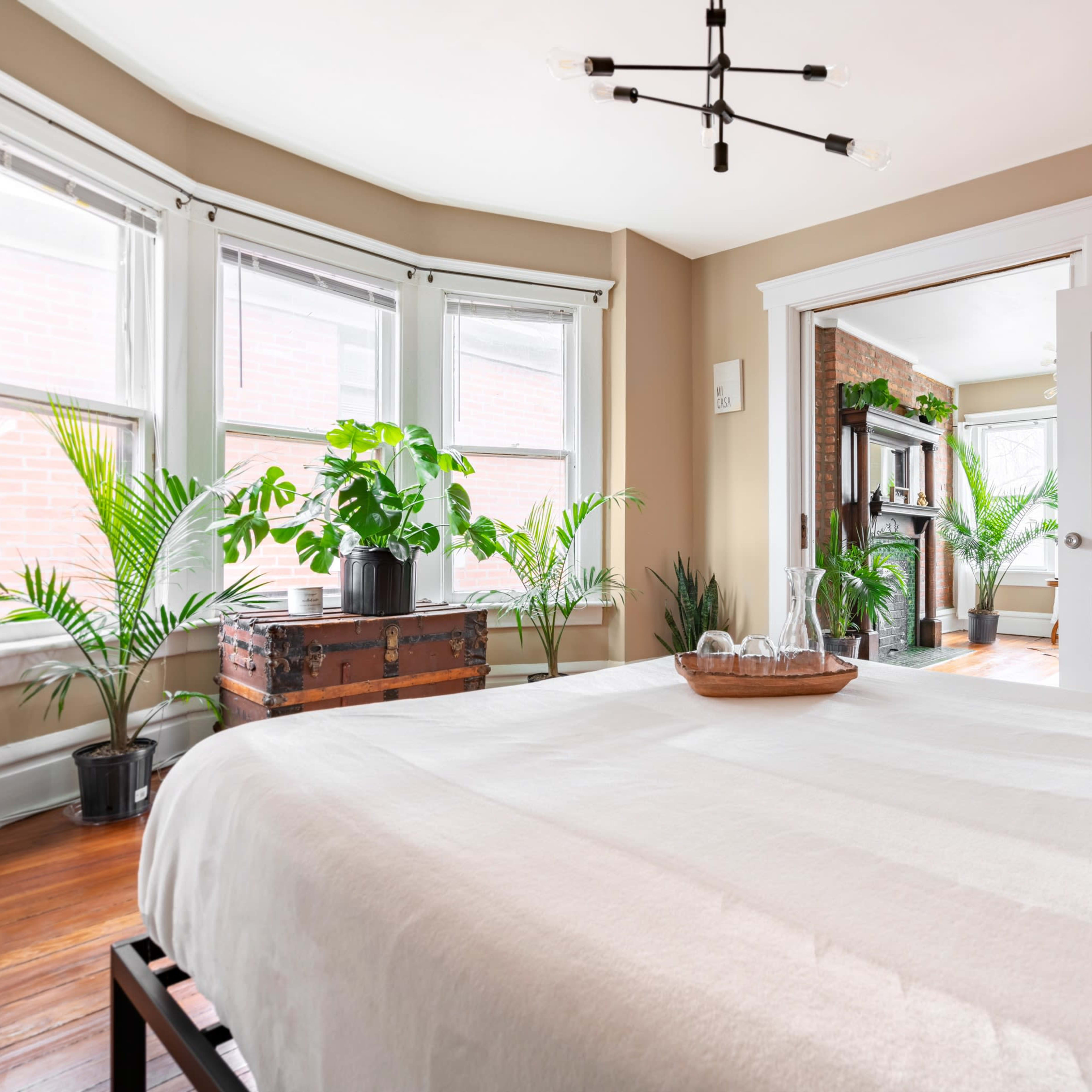 A well-lit bedroom features a bed with a white coverlet, large windows, and potted plants, leading to an adjoining room.