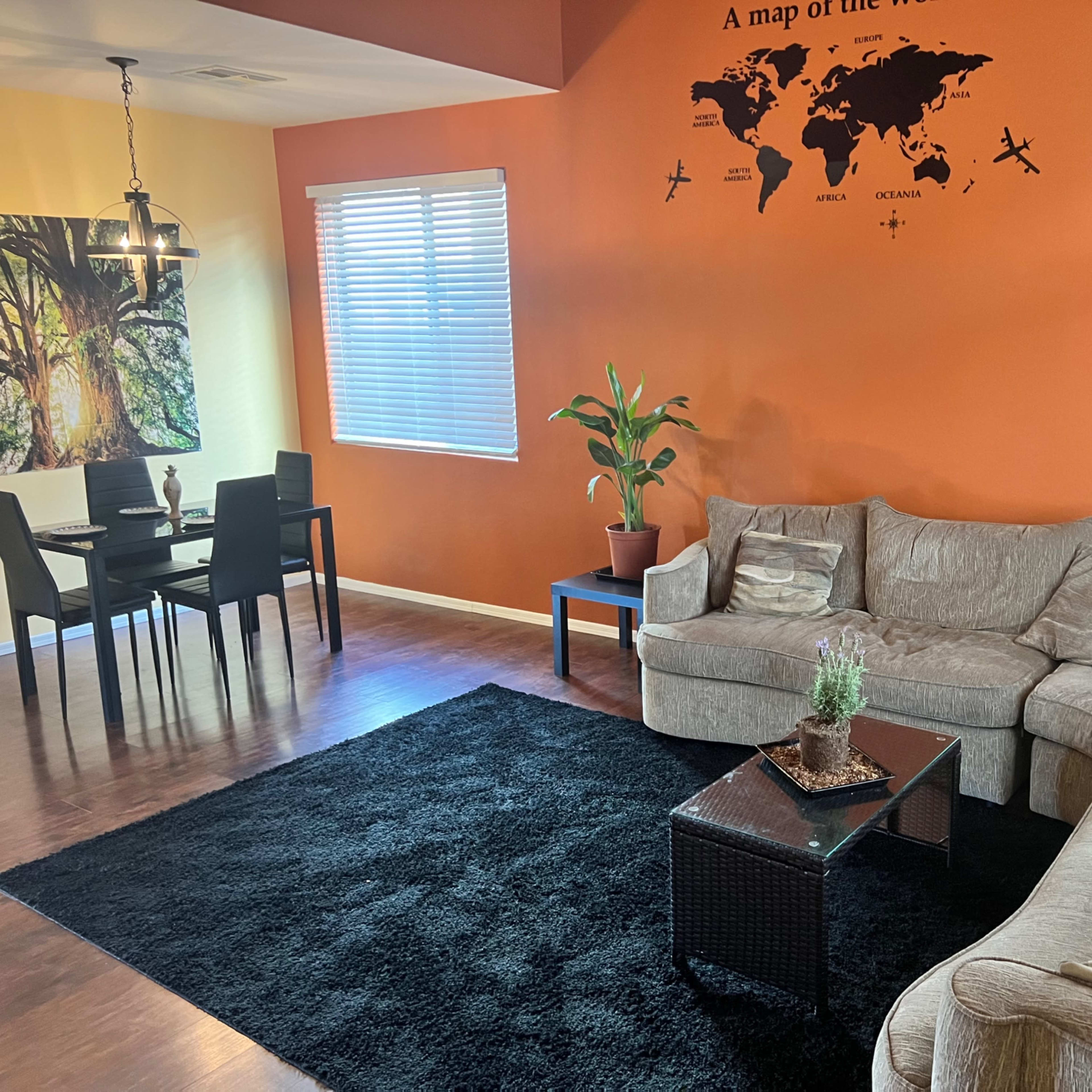 The room features a sectional sofa, a black area rug, a small coffee table, a potted plant, and a dining area with a table and chairs against a brightly colored wall with a world map design.
