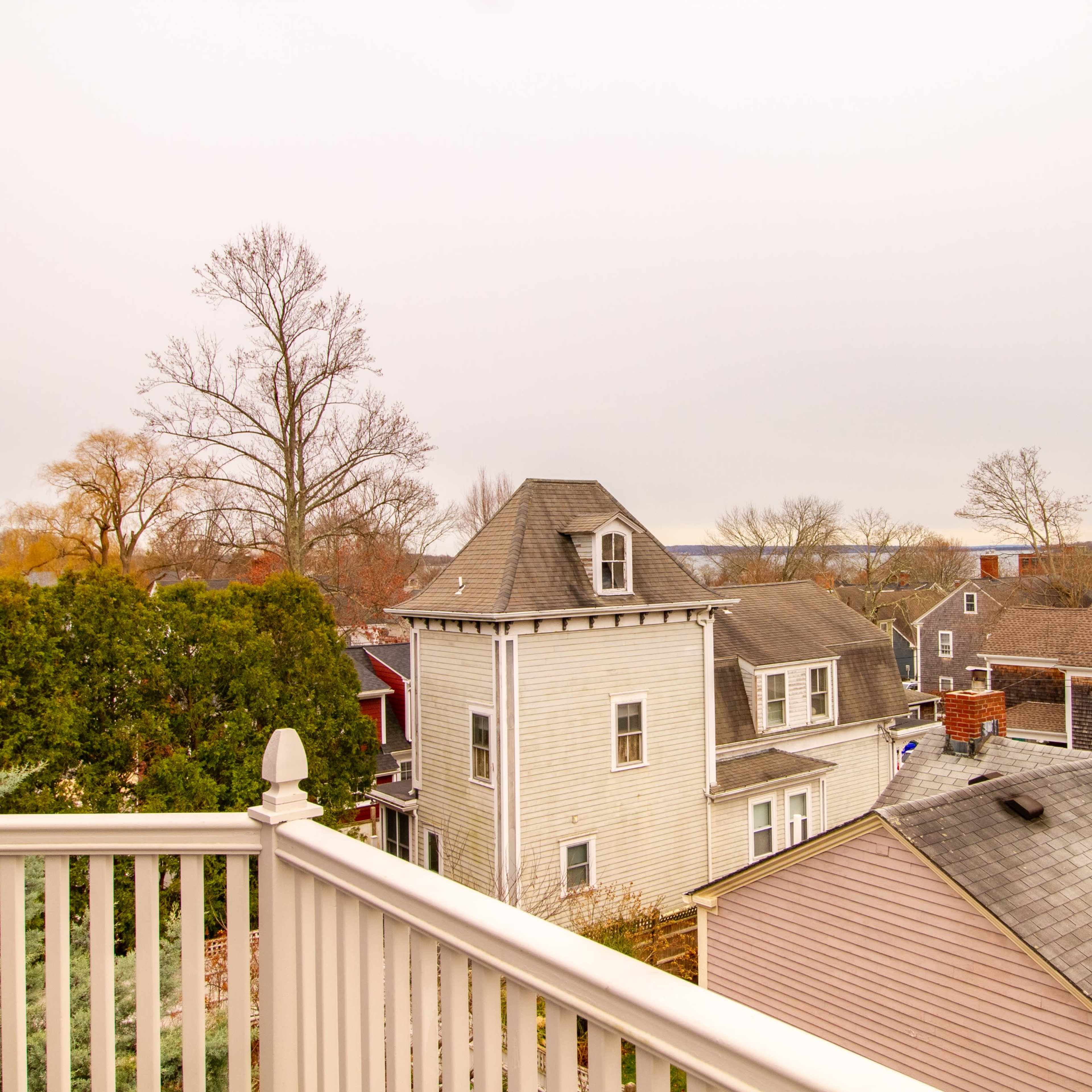 A view from a balcony overlooking a neighborhood with various houses, trees, and a distant body of water under a cloudy sky.