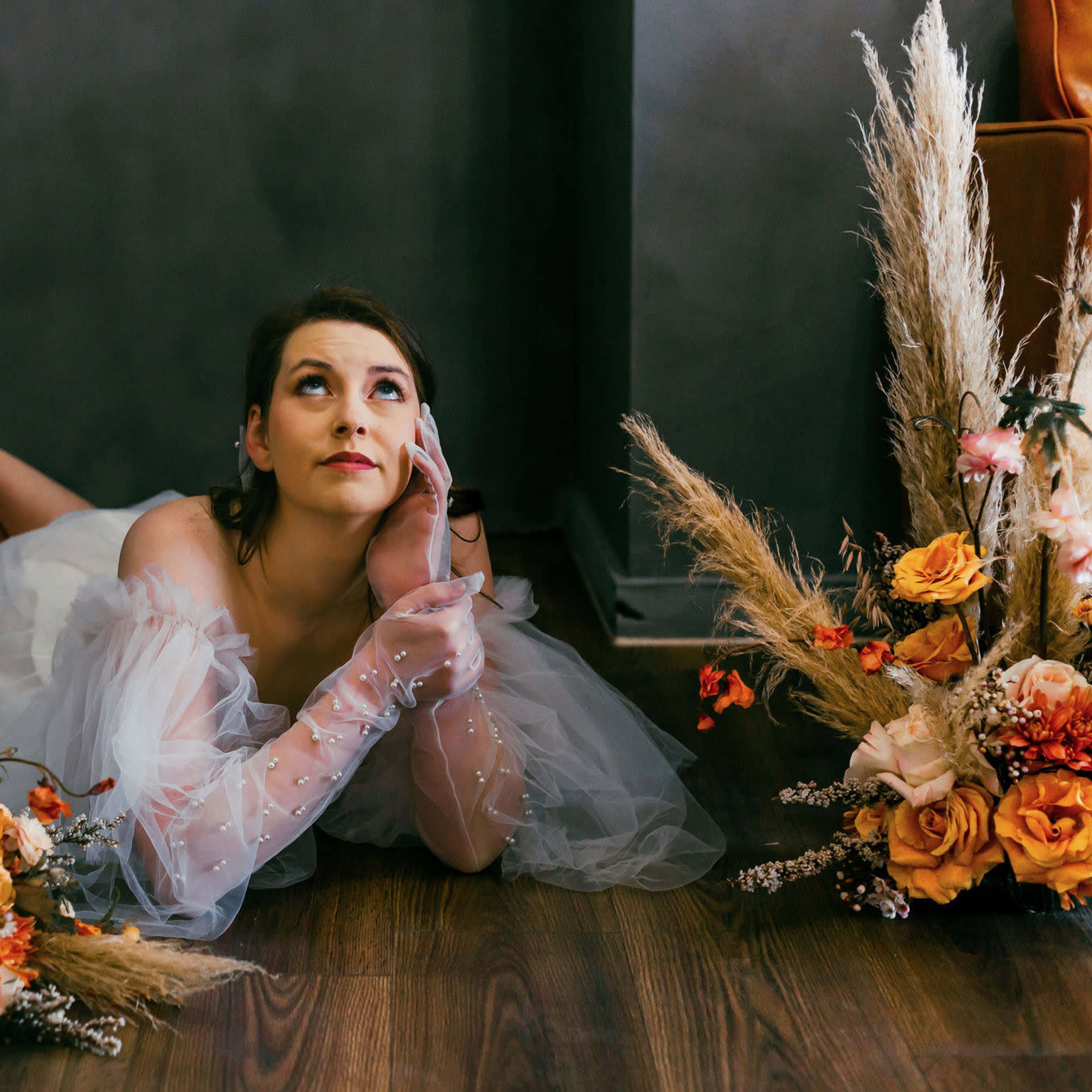 A bride in a white dress lies on a wooden floor, resting her chin on her hand, surrounded by floral arrangements.