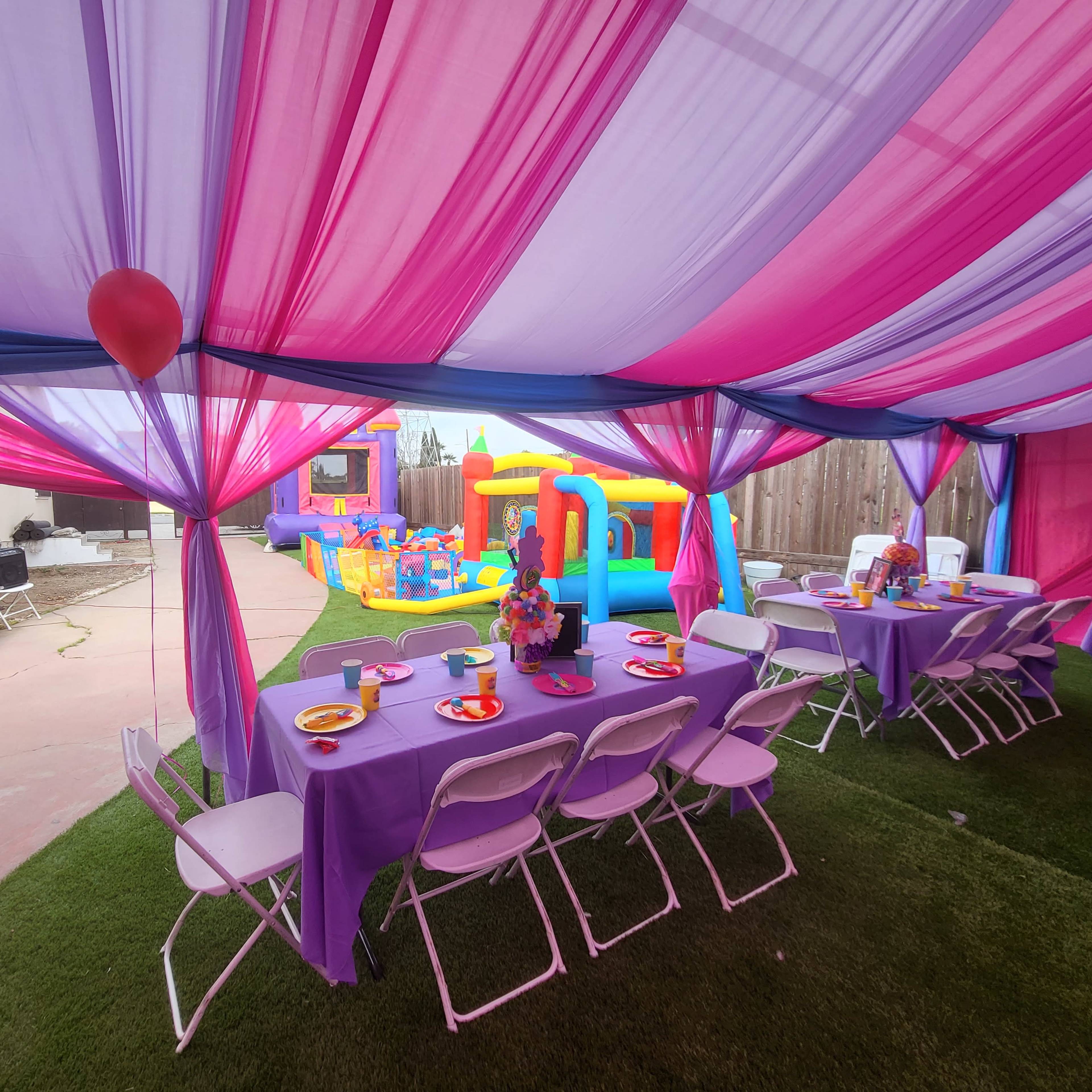 A tent decorated with pink and purple drapes houses tables set for a party, with a bouncy castle visible in the backyard.