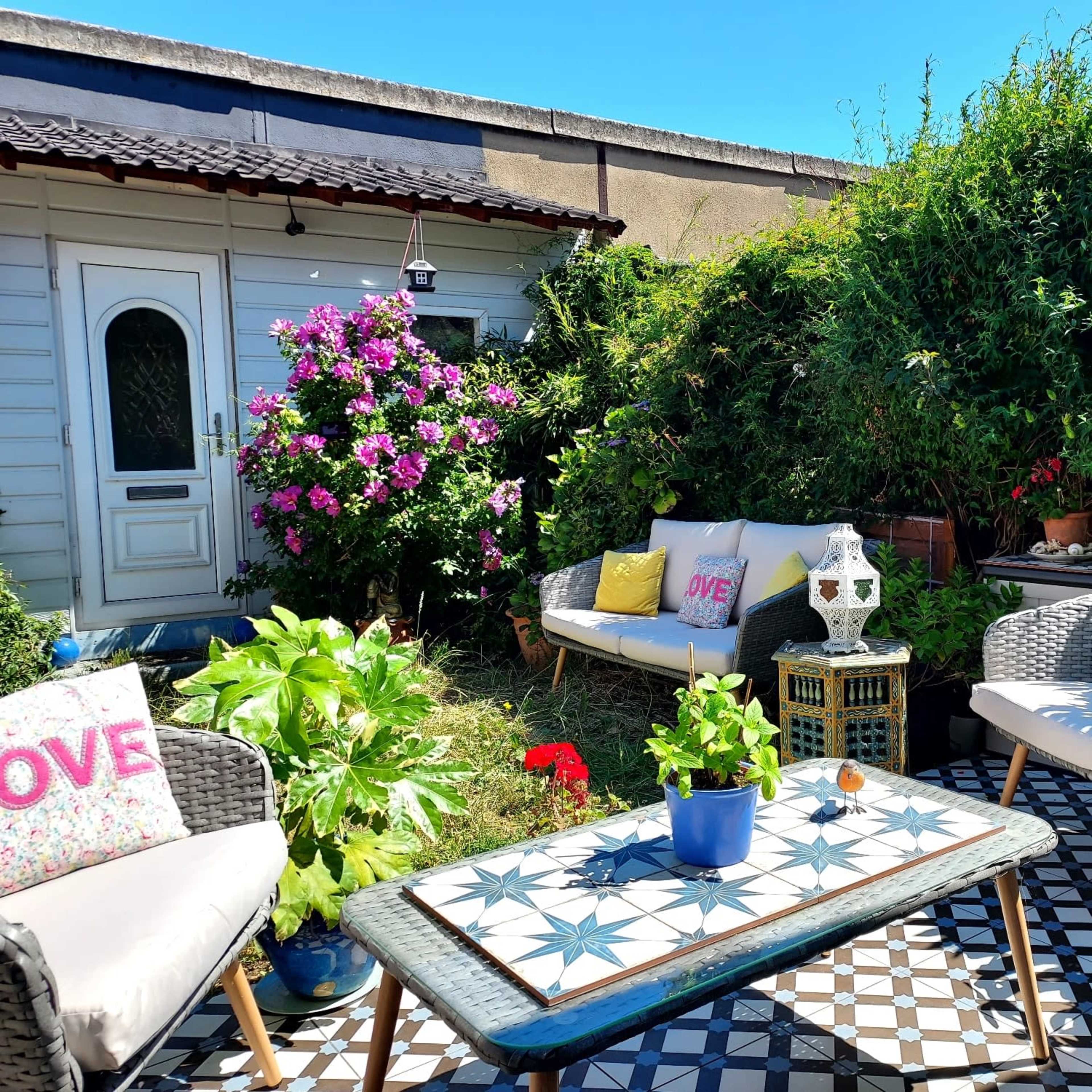 A cozy outdoor seating area features a table with a patterned design, surrounded by lush greenery and colorful flowers, alongside a small house.
