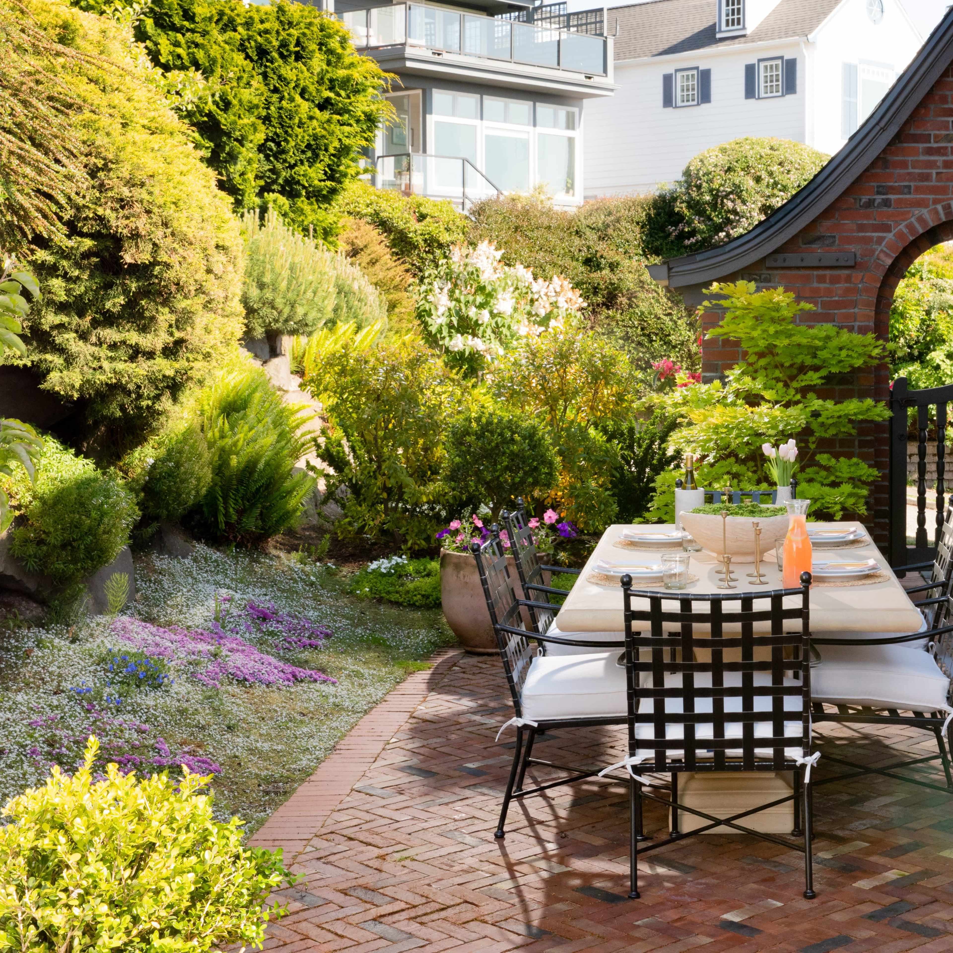 A brick patio features a dining table set for outdoor meals, surrounded by neatly arranged flower beds and greenery.