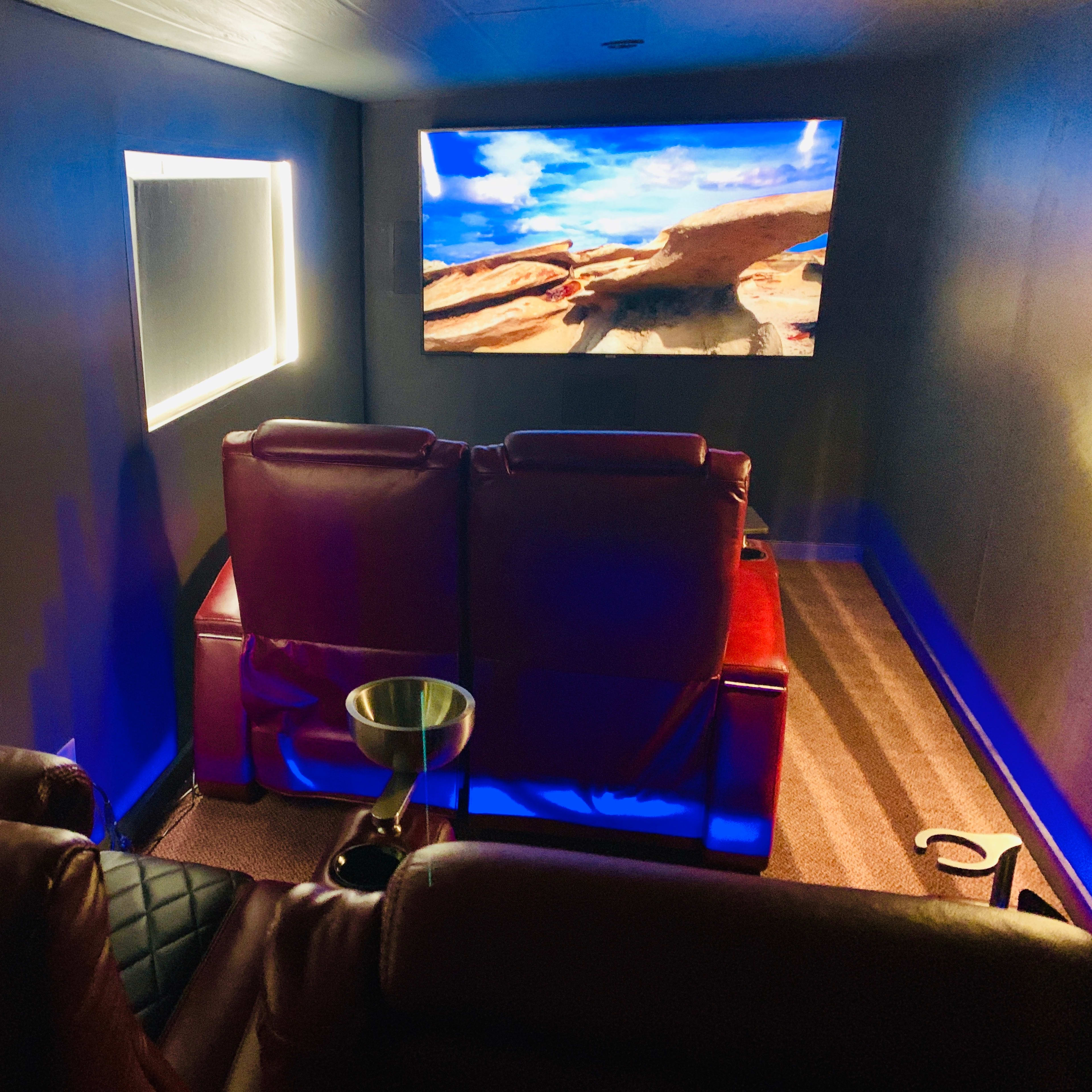 A dimly lit home theater features two red recliner chairs facing a large screen showing a scenic landscape.