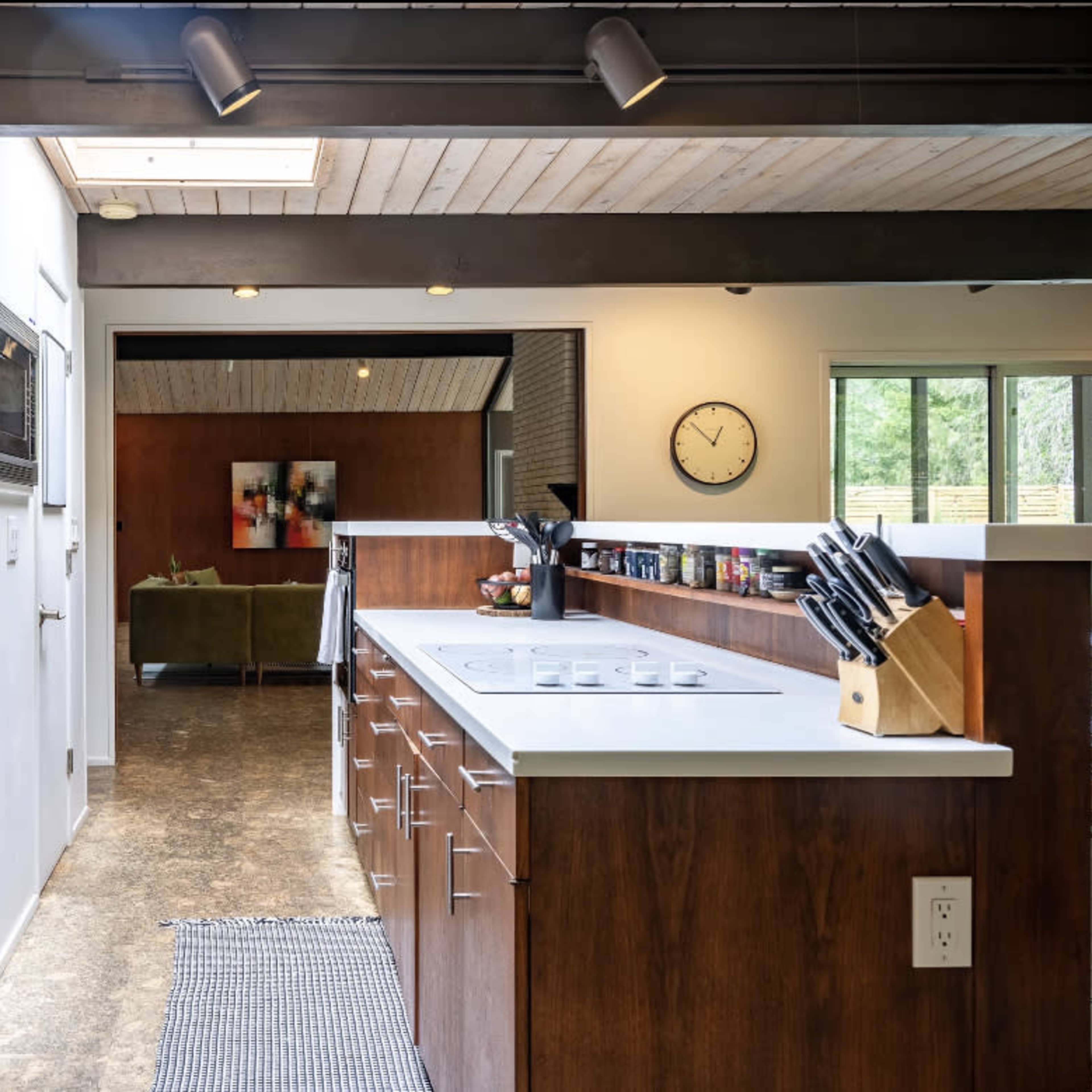 A modern kitchen features wooden cabinetry, a white countertop, and a mosaic tile backsplash, with an open view to a living area.