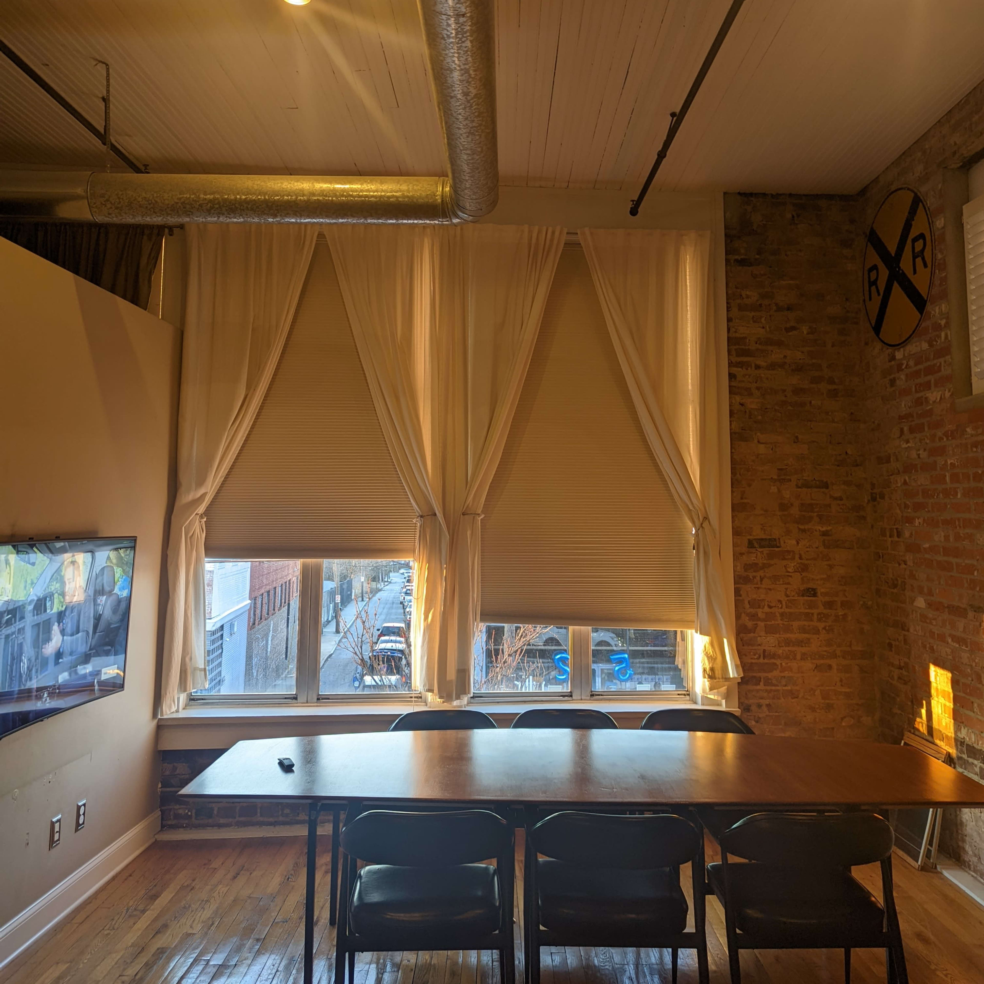 A conference room features a long wooden table with black chairs, large windows dressed in white curtains, and exposed brick walls.