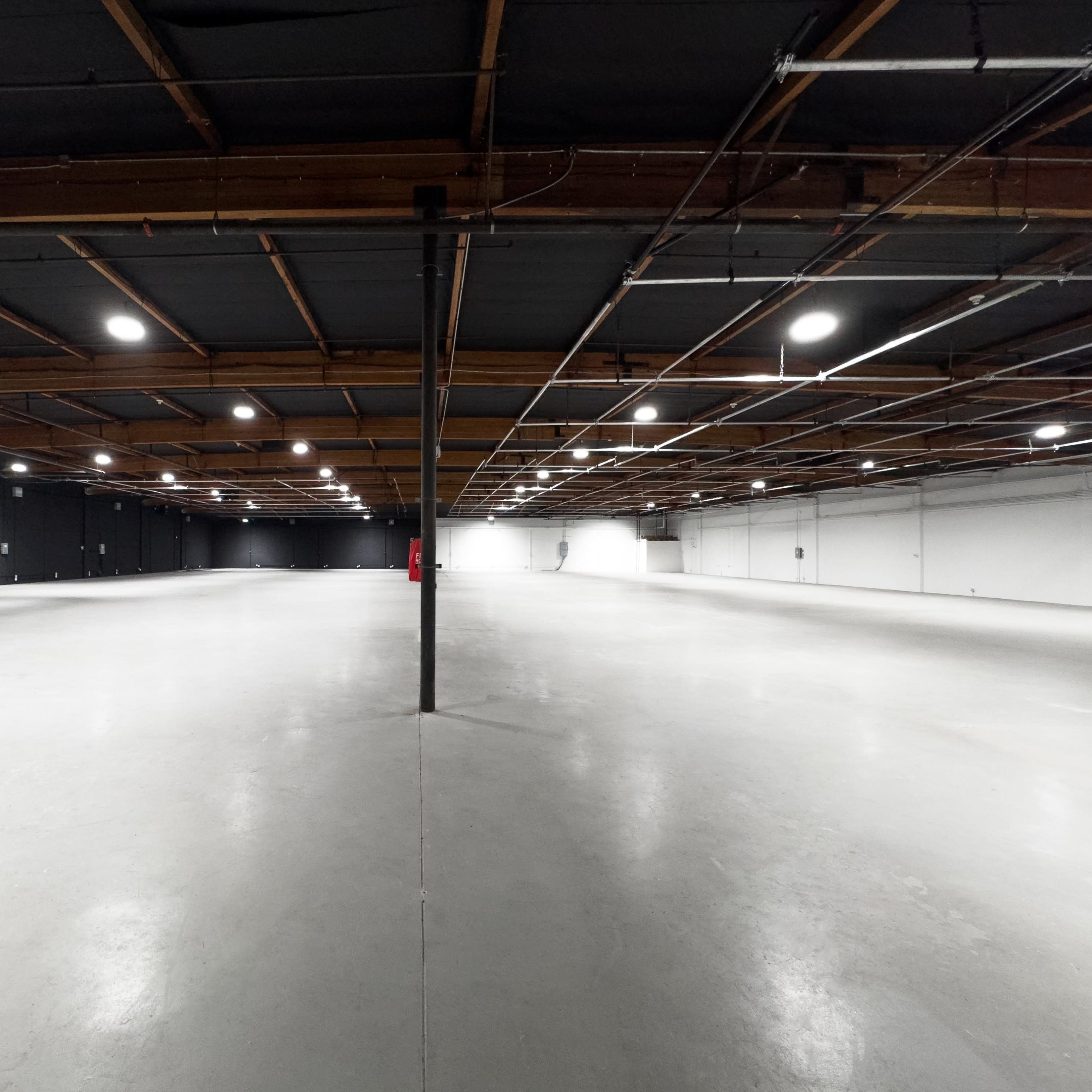 A spacious industrial warehouse features concrete flooring and exposed wooden beams, with bright overhead lighting illuminating the empty interior.