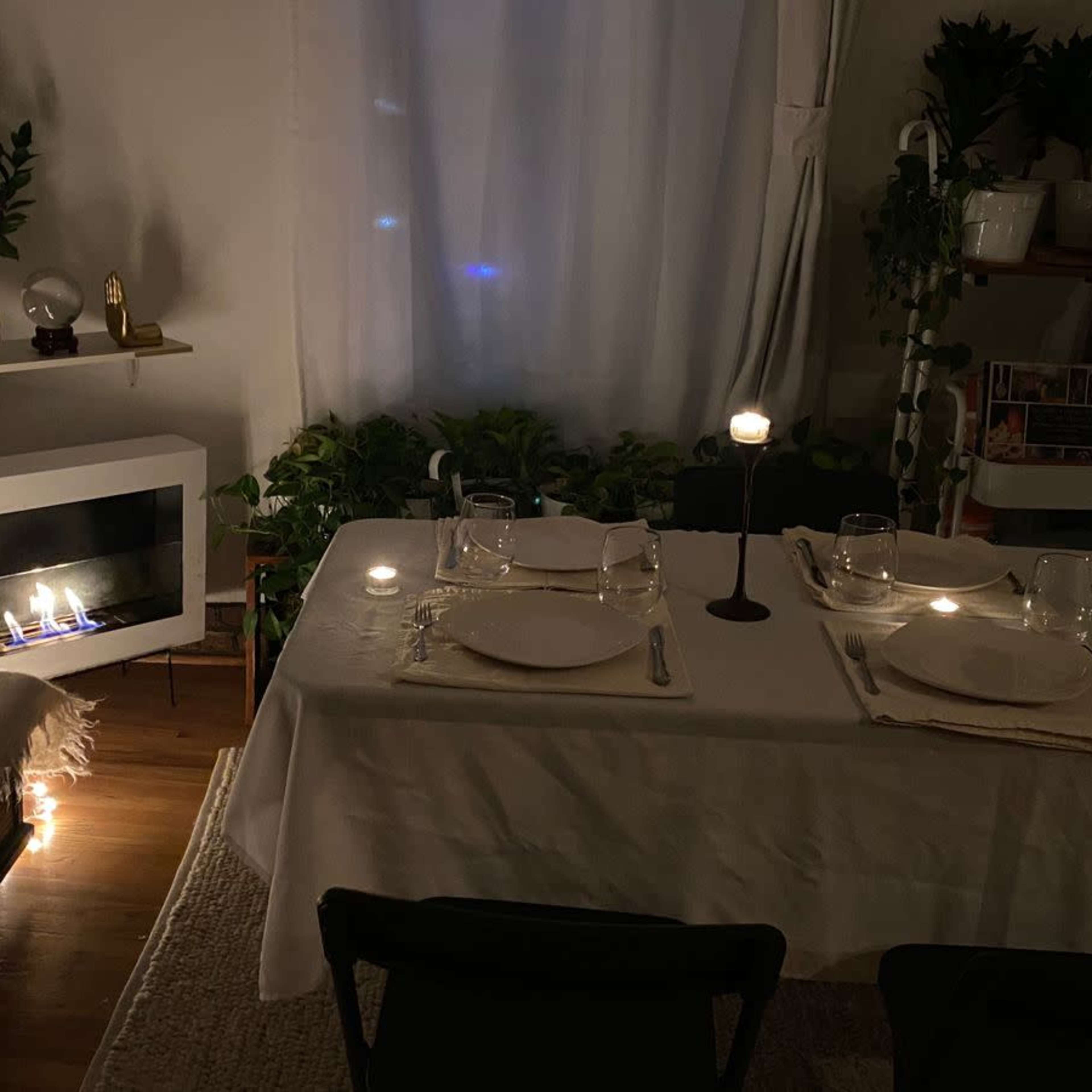 The scene depicts a dimly lit dining area with a neatly set table, surrounded by plants and featuring a decorative fireplace with candles.