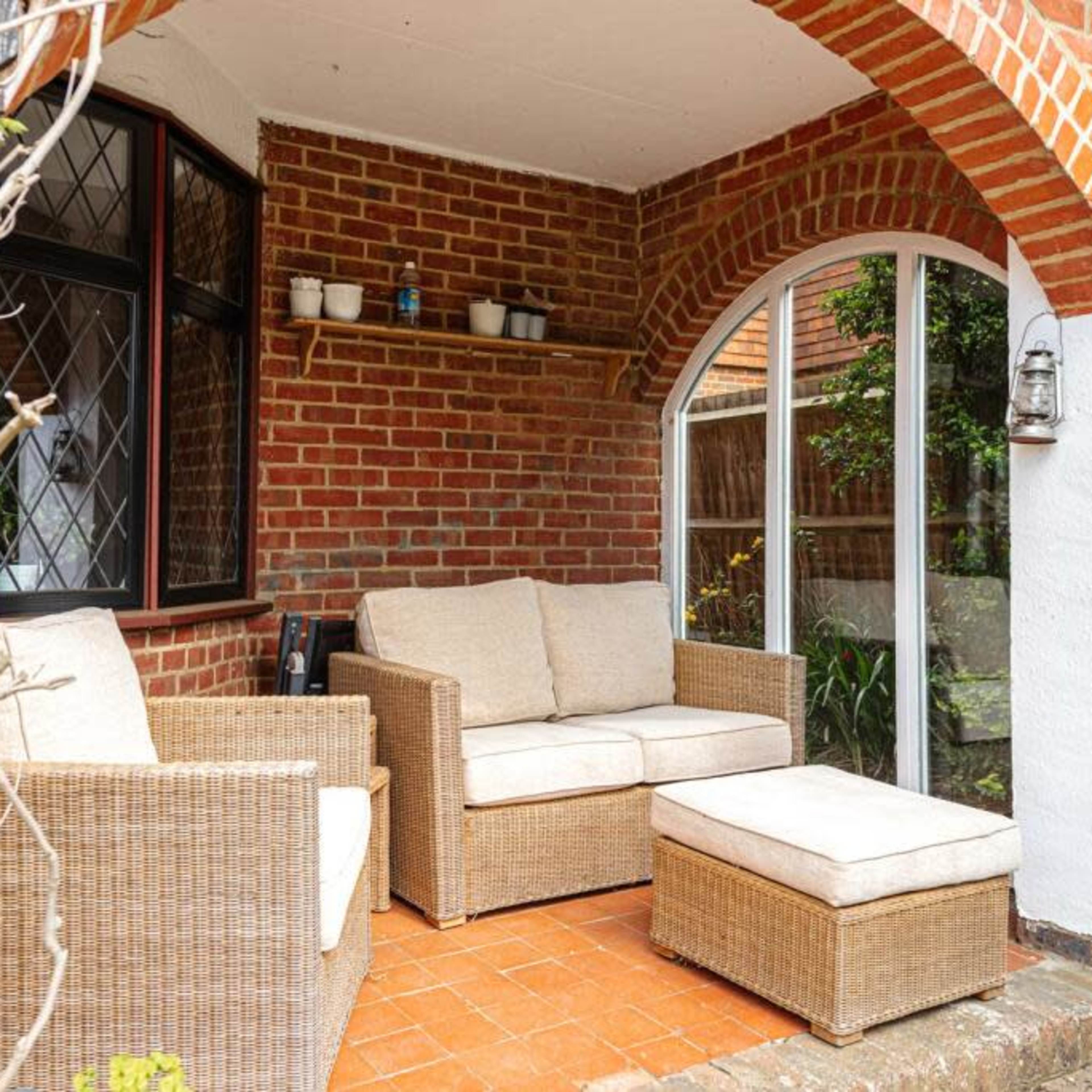A cozy outdoor seating area with rattan furniture, including a sofa and an ottoman, set against a brick wall under an archway.