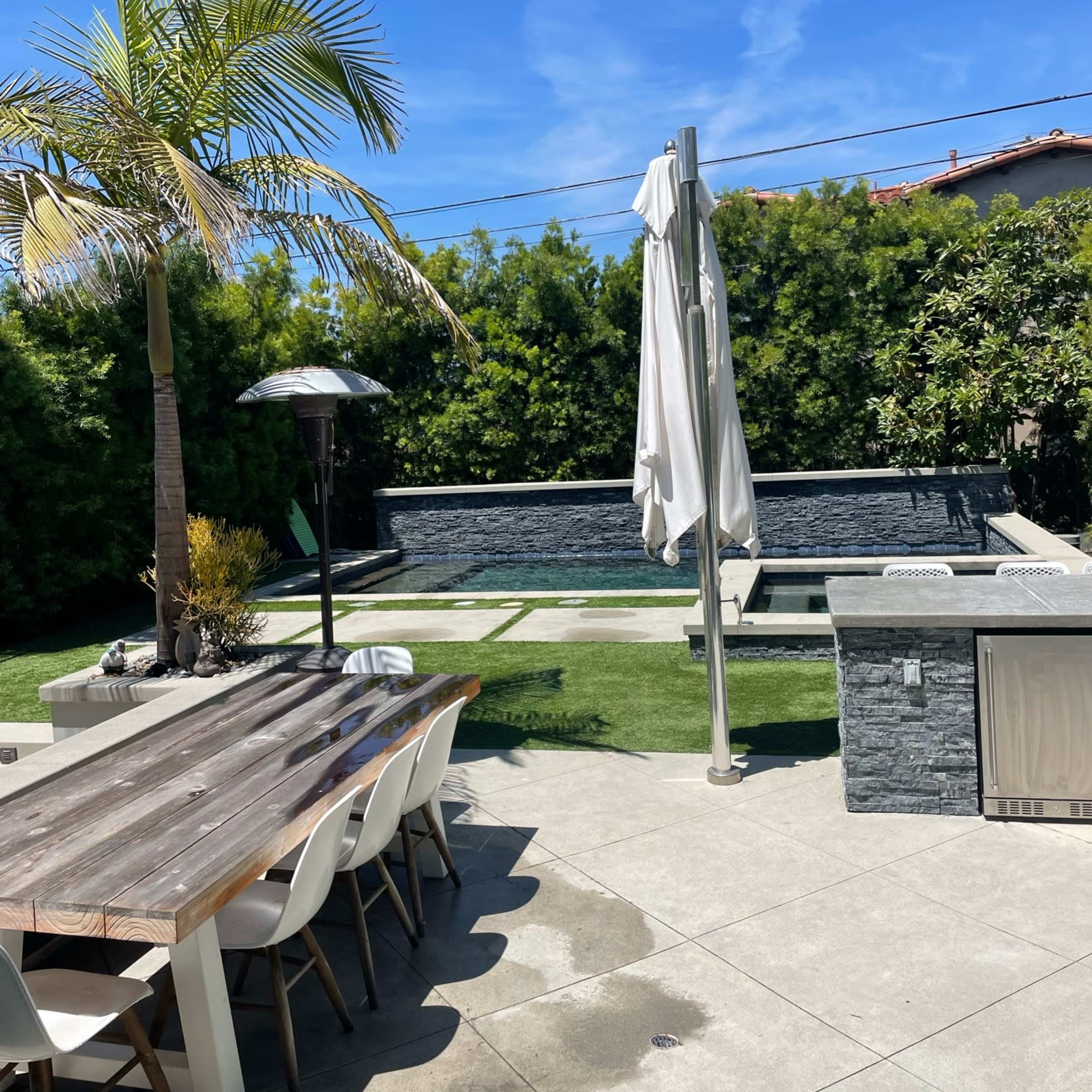 A backyard scene features a dining table and chairs, a pool surrounded by greenery, and a palm tree under a clear blue sky.