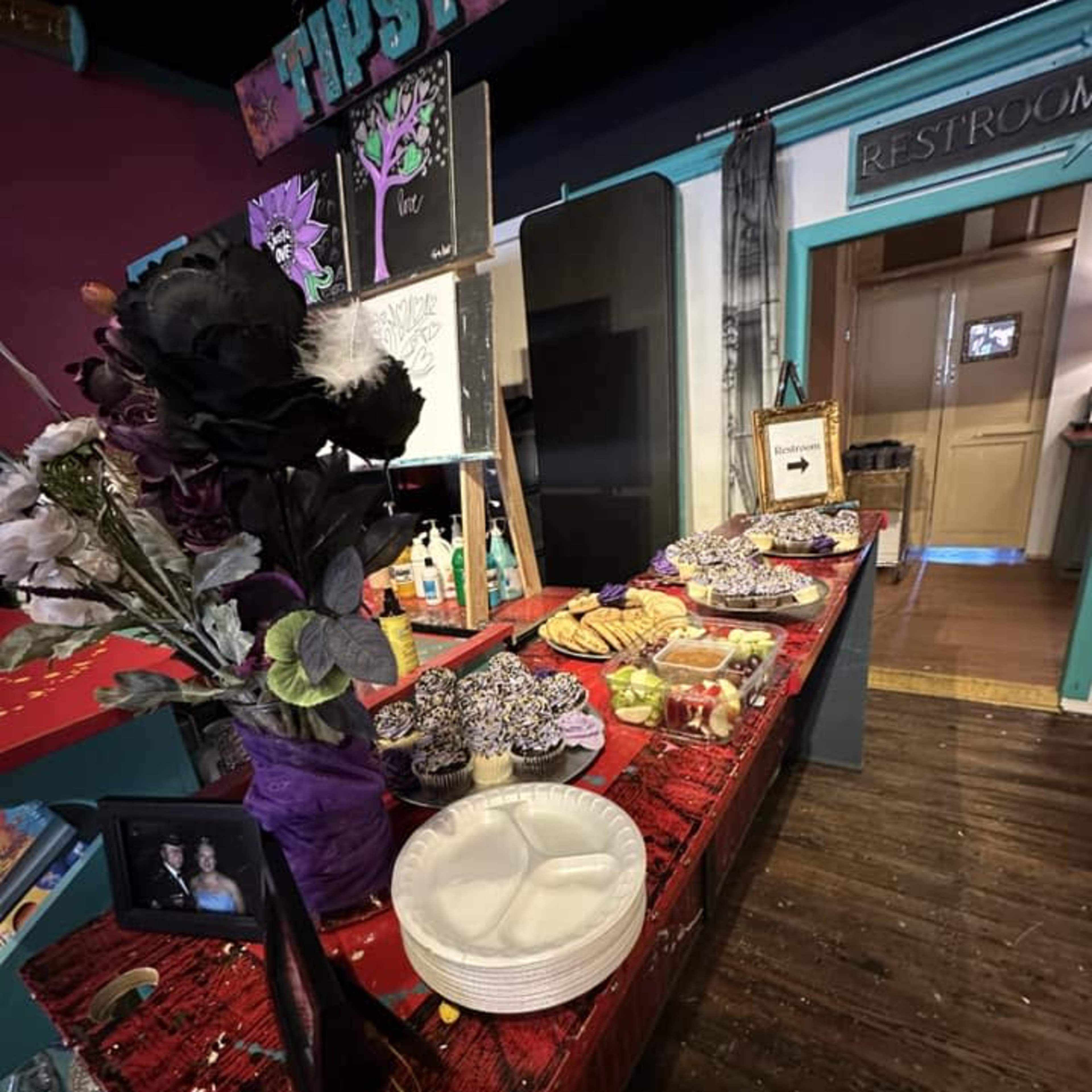 A table covered with food items, plates, and a decorative flower arrangement inside a colorful venue featuring a restroom sign in the background.