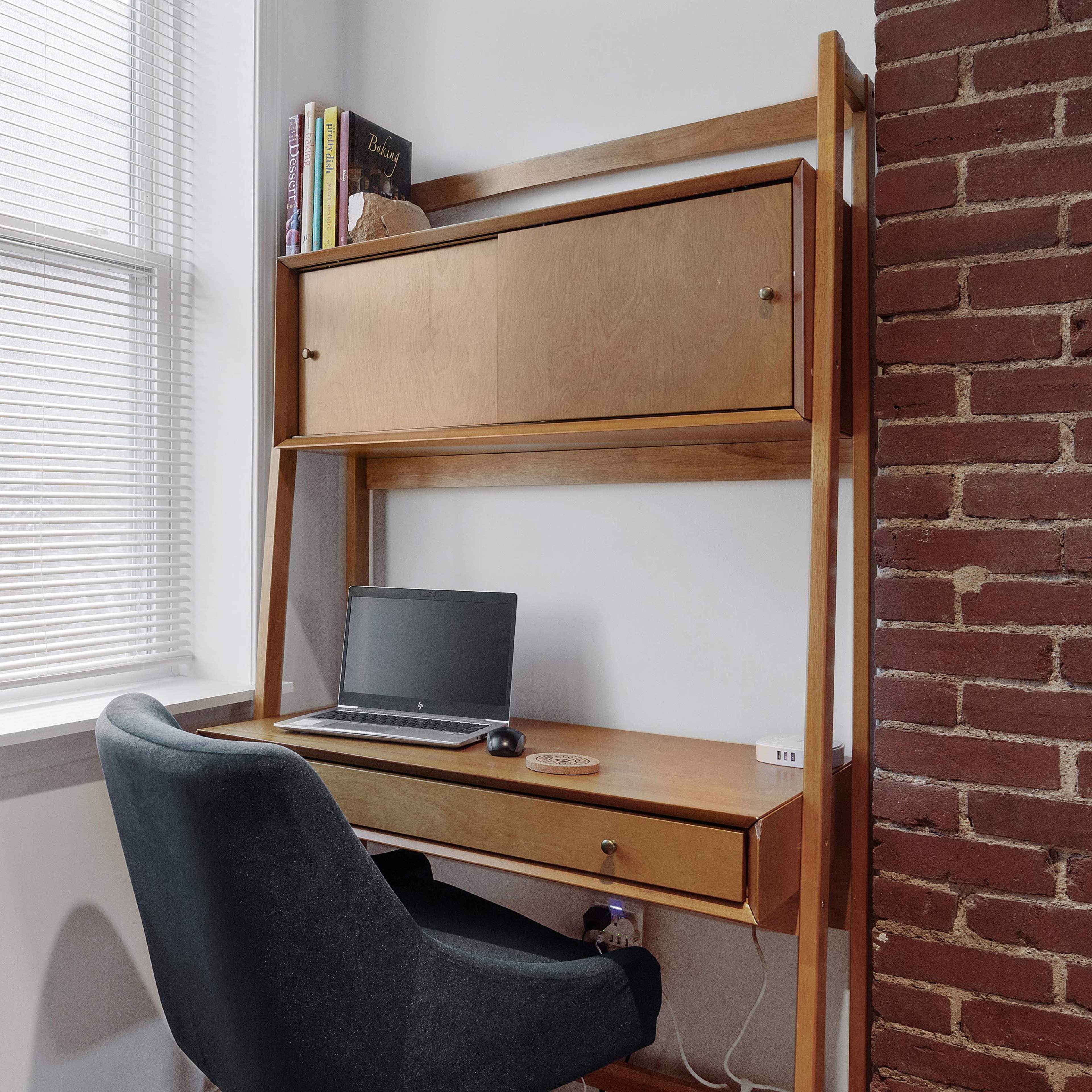 A wooden desk with a laptop sits beside a brick wall, illuminated by natural light from a nearby window with blinds.