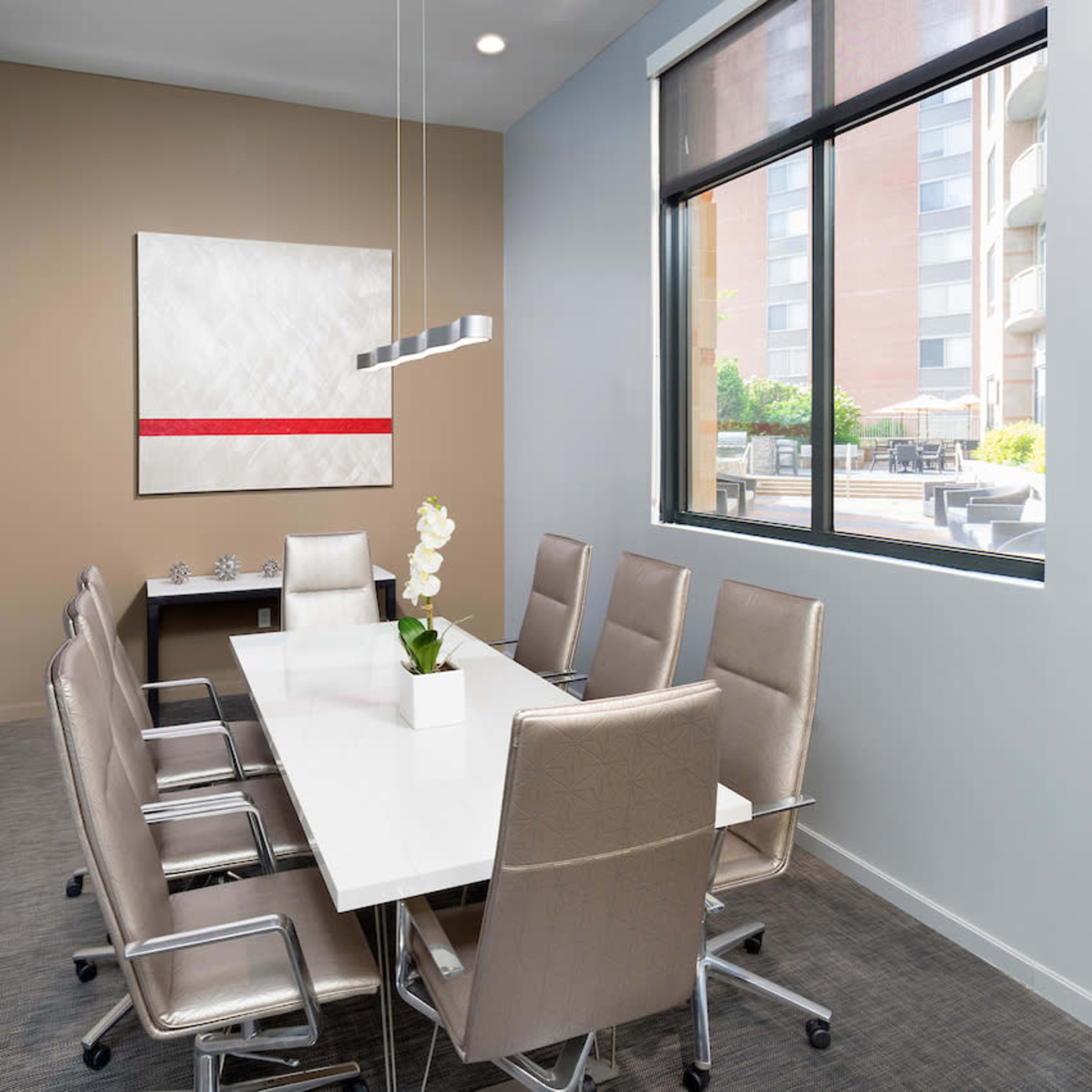 A modern conference room features a long white table surrounded by six silver chairs, with a television mounted on the wall and a large window providing natural light.