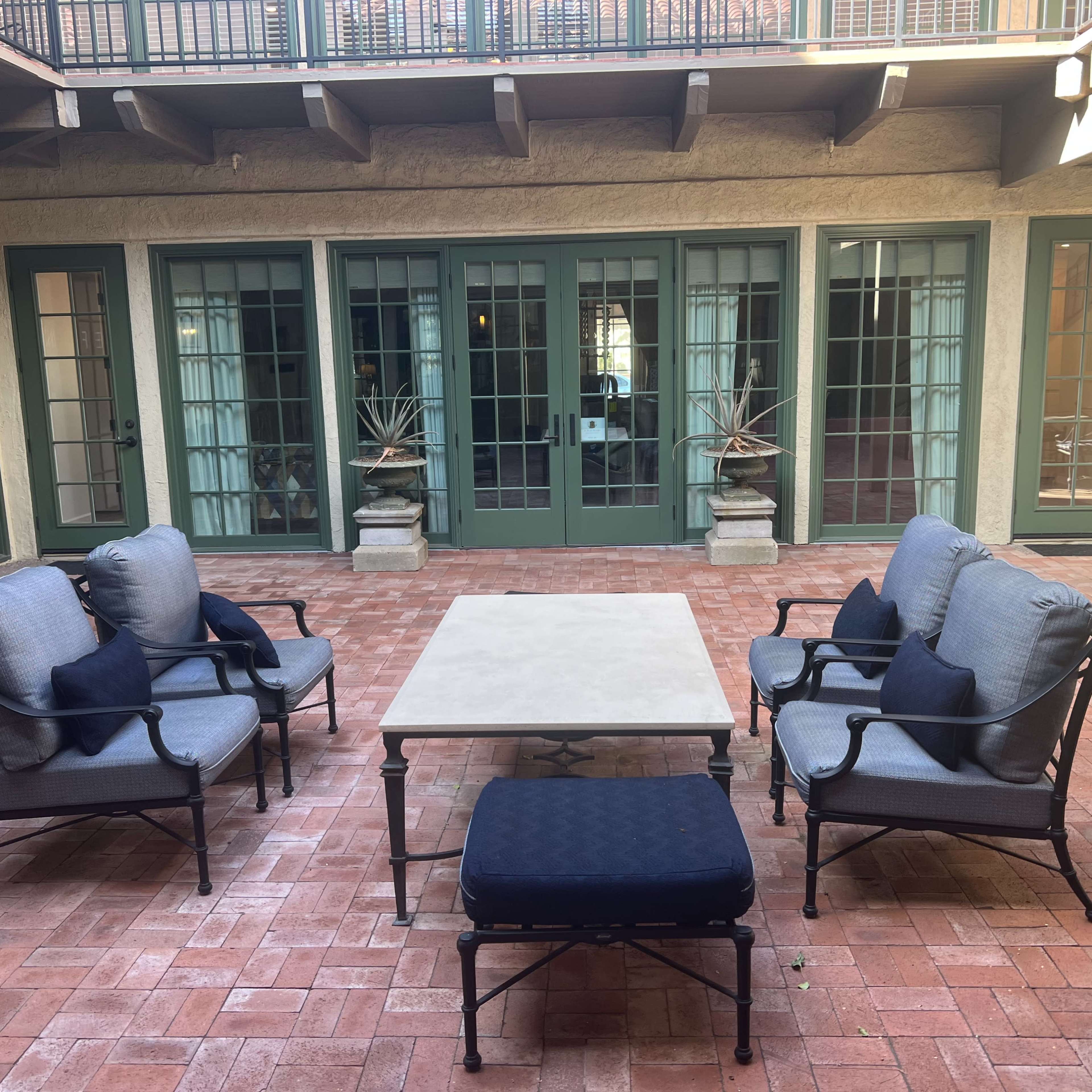 A courtyard features a set of four armchairs and a coffee table arranged on a brick floor, with large green-paneled windows in the background.