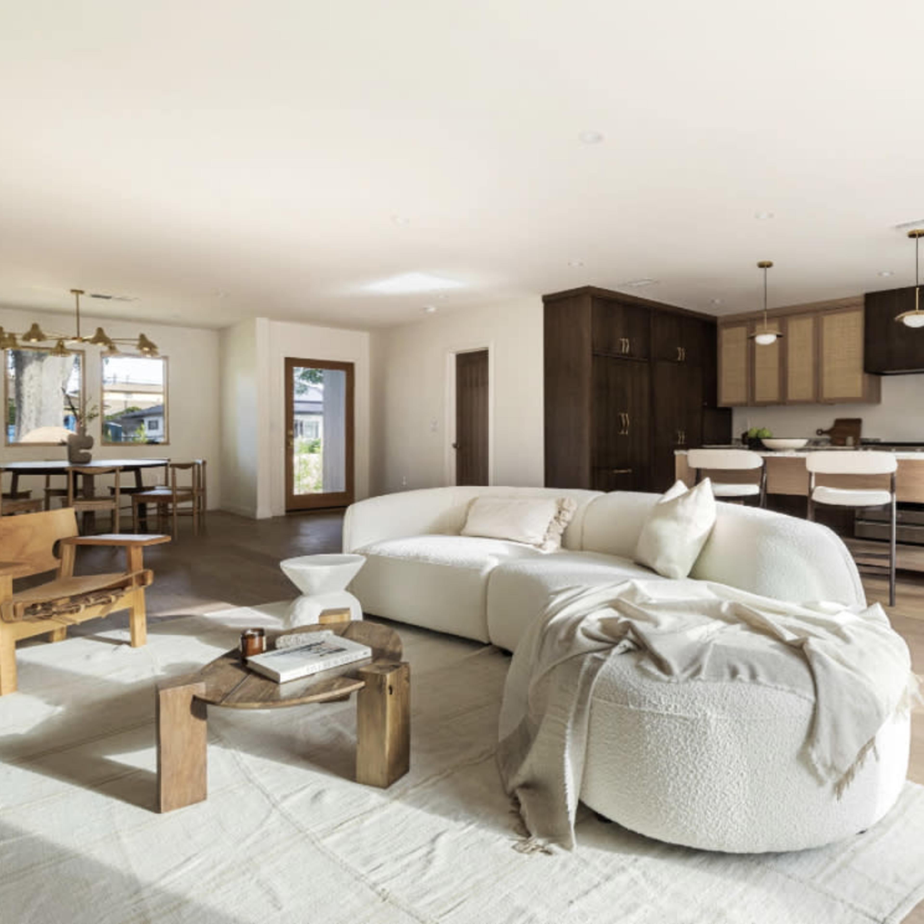 A spacious living area features a curved sofa, a wooden coffee table, and leads into a modern kitchen with light-colored cabinetry and a dining table nearby.