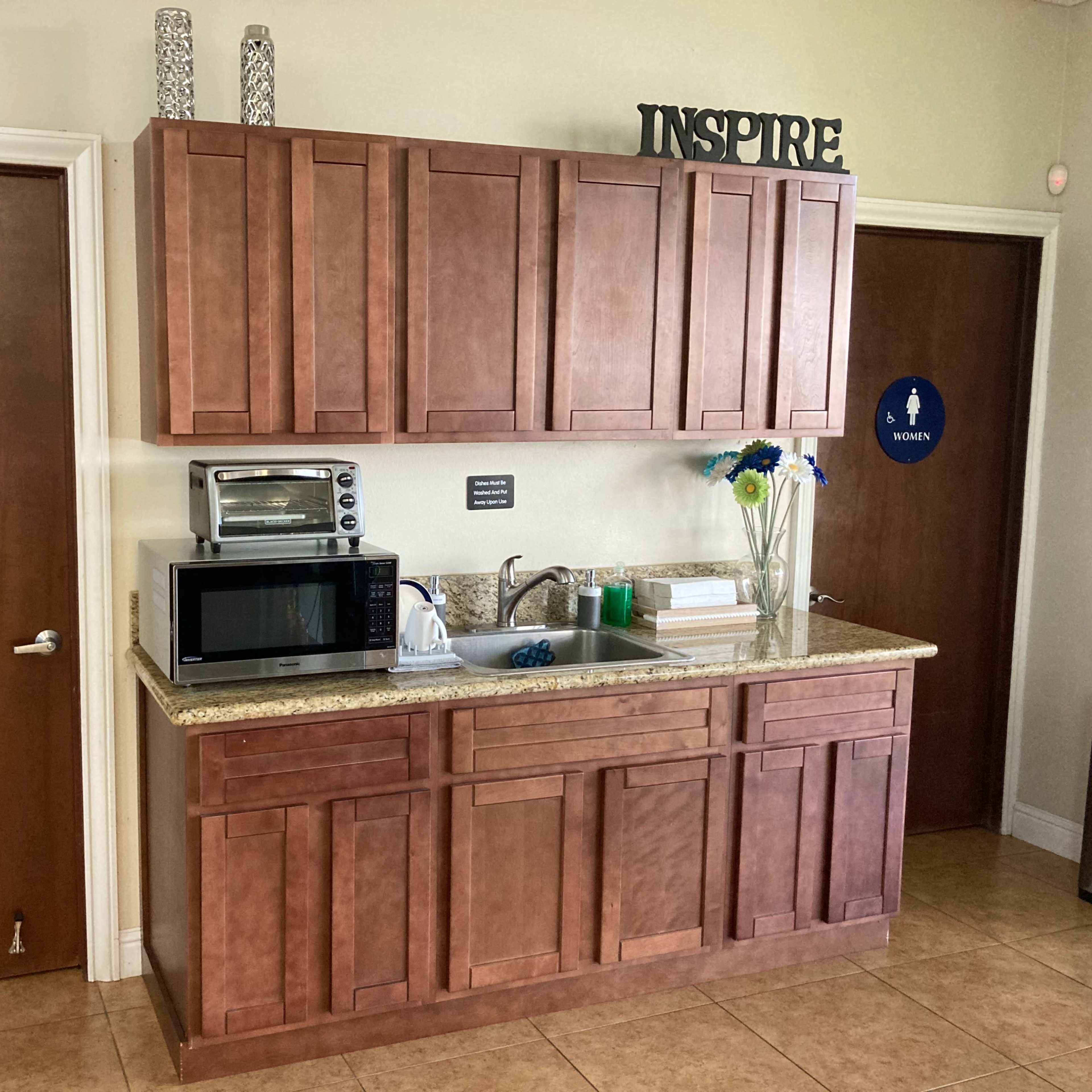 A kitchenette with wooden cabinets, a microwave, and a granite countertop, alongside two restroom doors labeled "MEN" and "WOMEN."