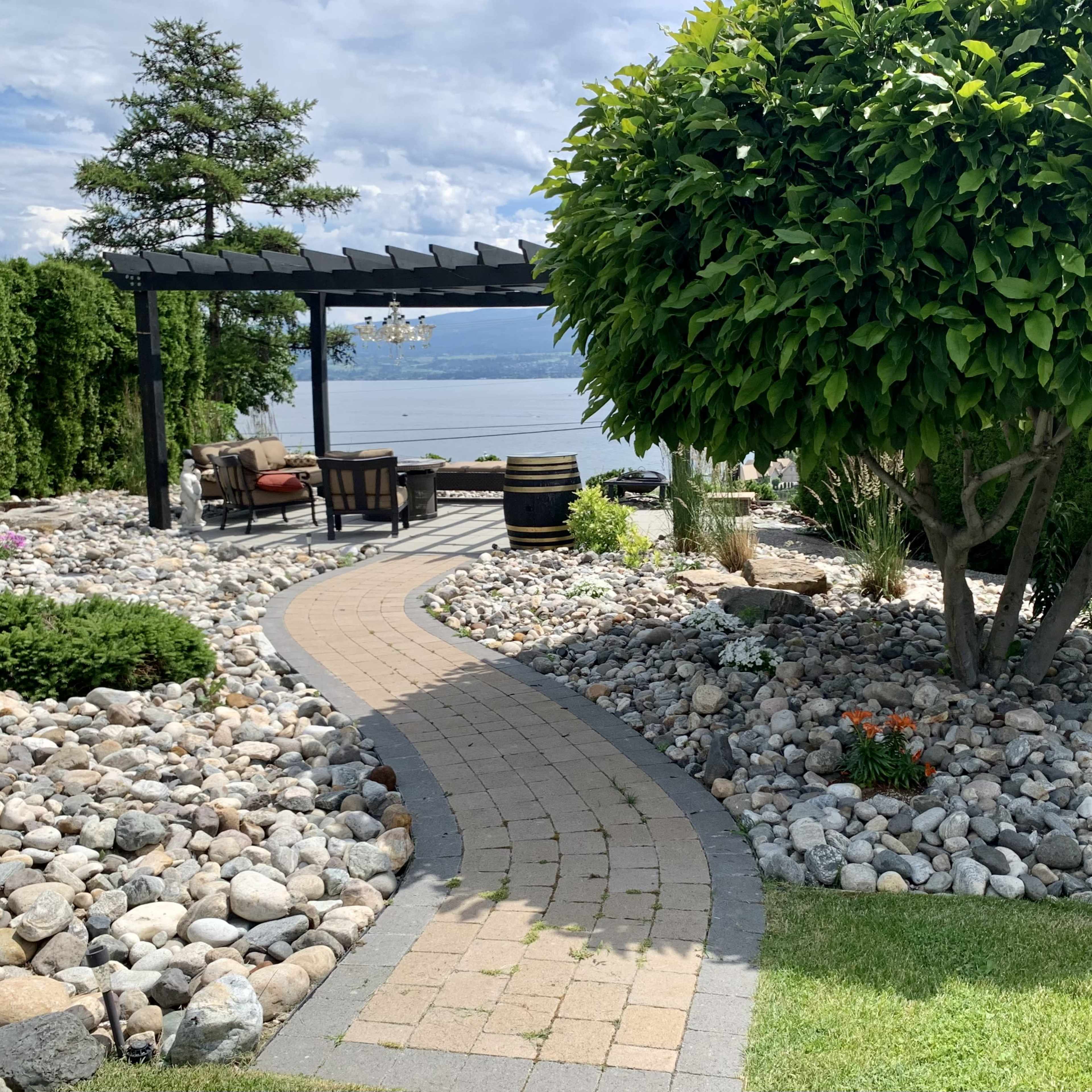 A winding stone pathway leads through a landscaped garden with rocks, shrubs, and a seating area overlooking a body of water.